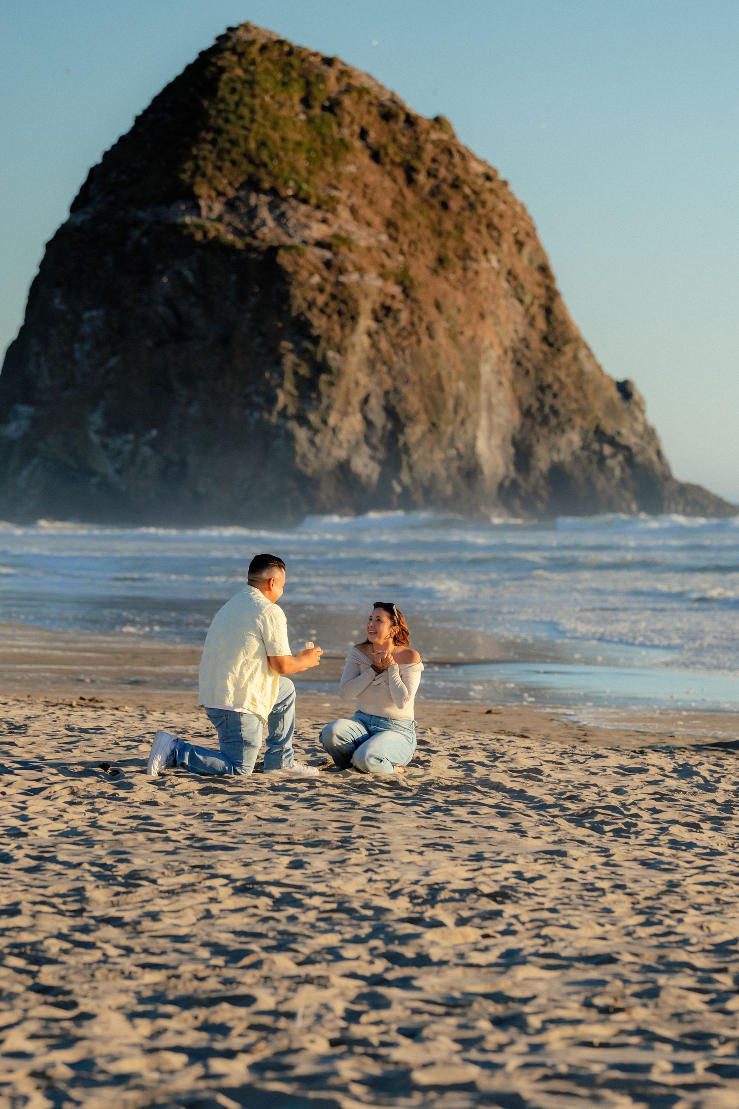 Man proposes to woman on sandy beach with iconic Haystack Rock at Cannon Beach, Oregon in the background.