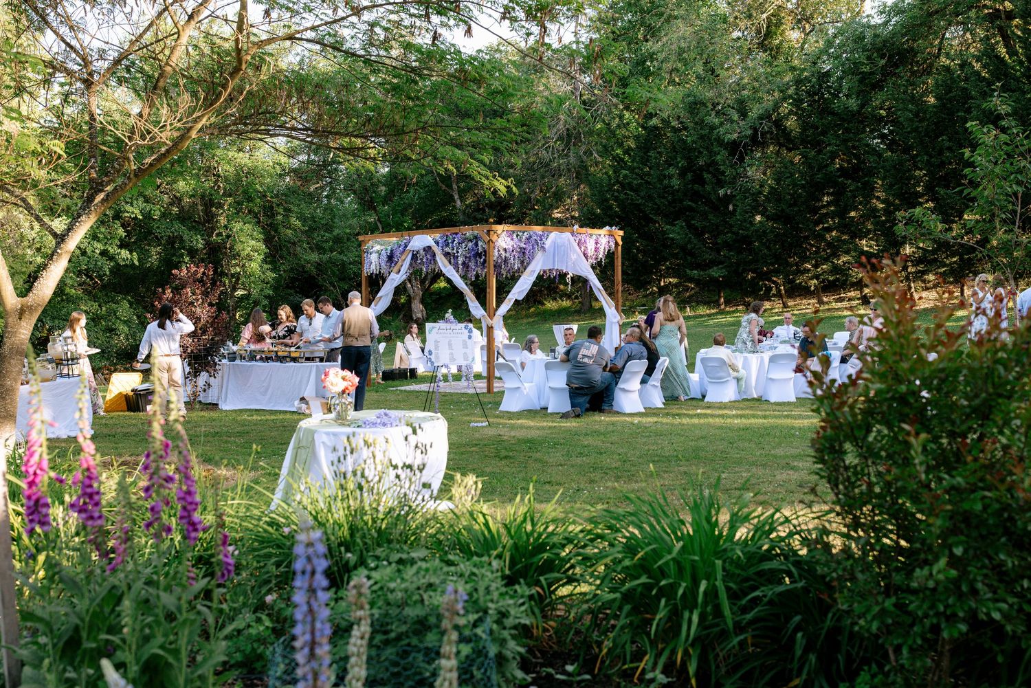 Elegant outdoor wedding reception setup with white tables and floral arch amid lush garden greenery.
