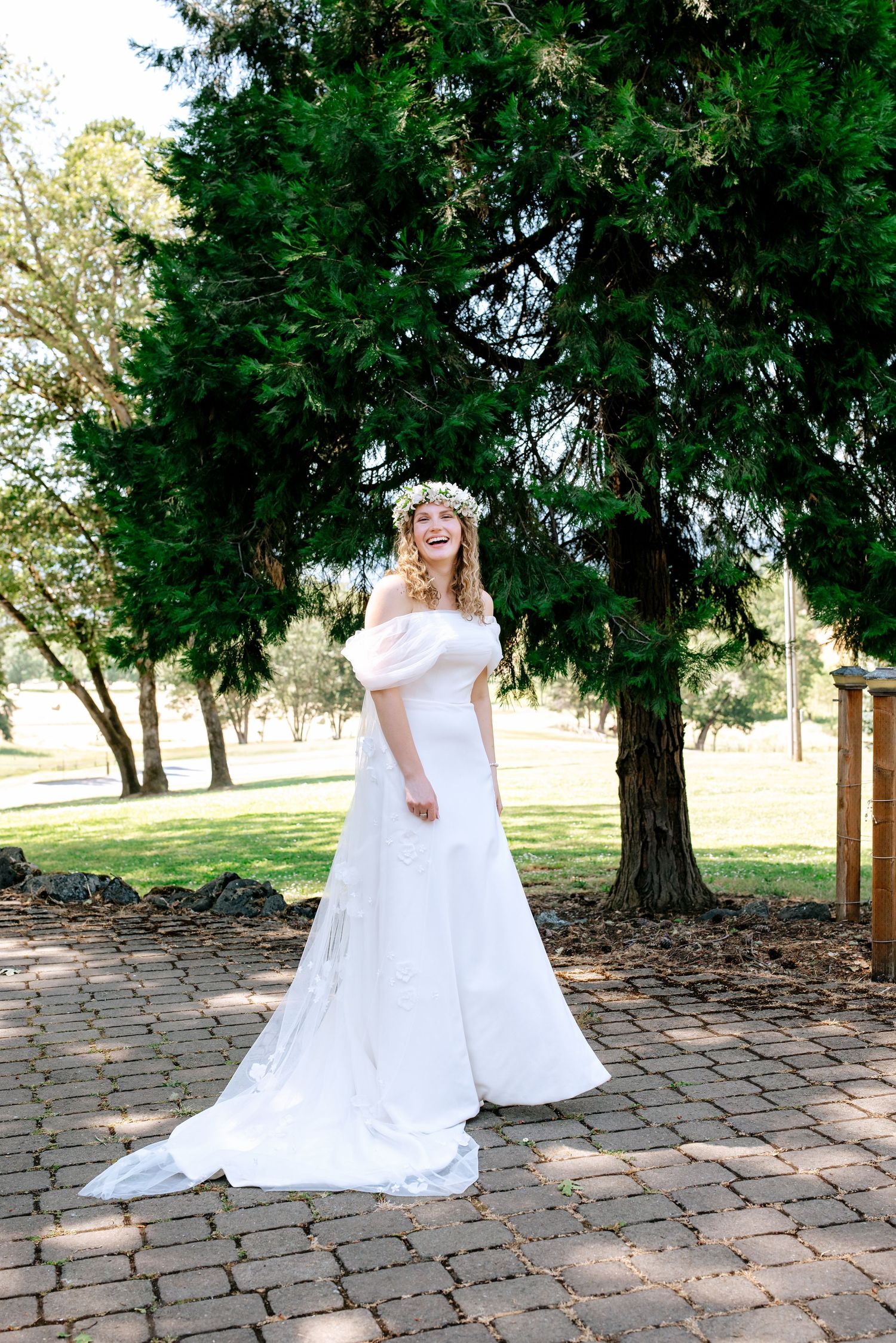 Bride in white off-shoulder gown and floral crown laughing joyfully in a sunlit park.