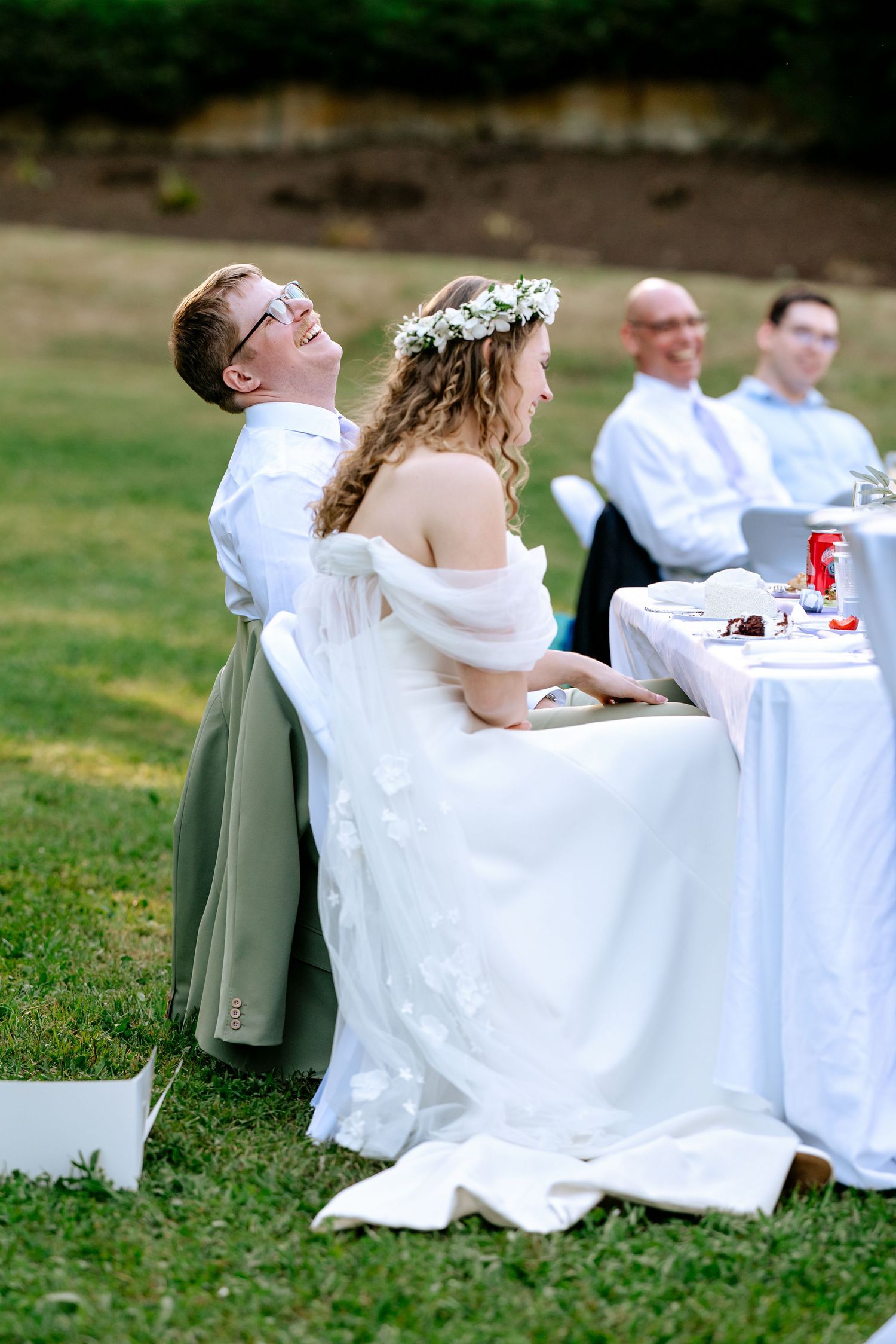 Bride in floral crown and groom in sage suit laugh together at outdoor wedding reception table.