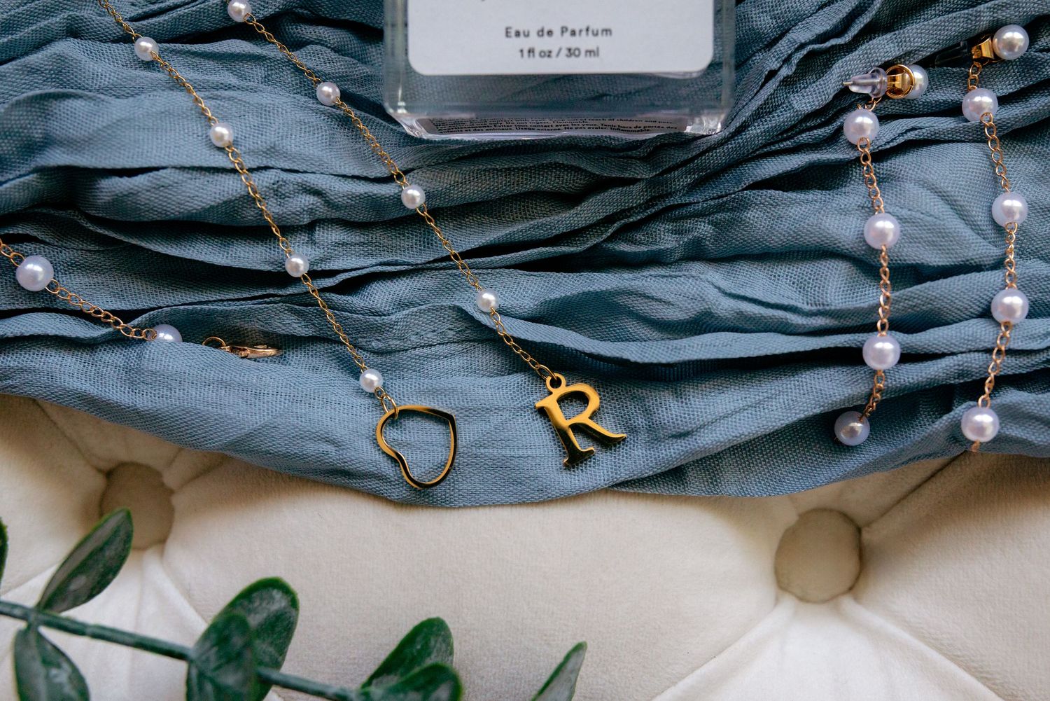Gold initial R pendant necklace with pearl chain and heart charm on blue silk fabric.