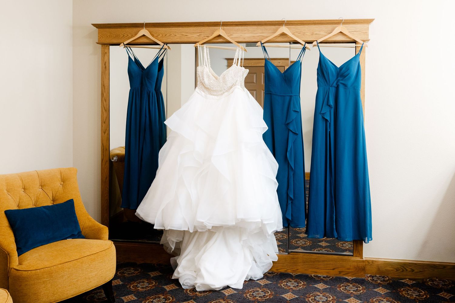 White wedding gown and blue bridesmaid dresses hanging on a wooden rack in an elegant room with yellow chair.