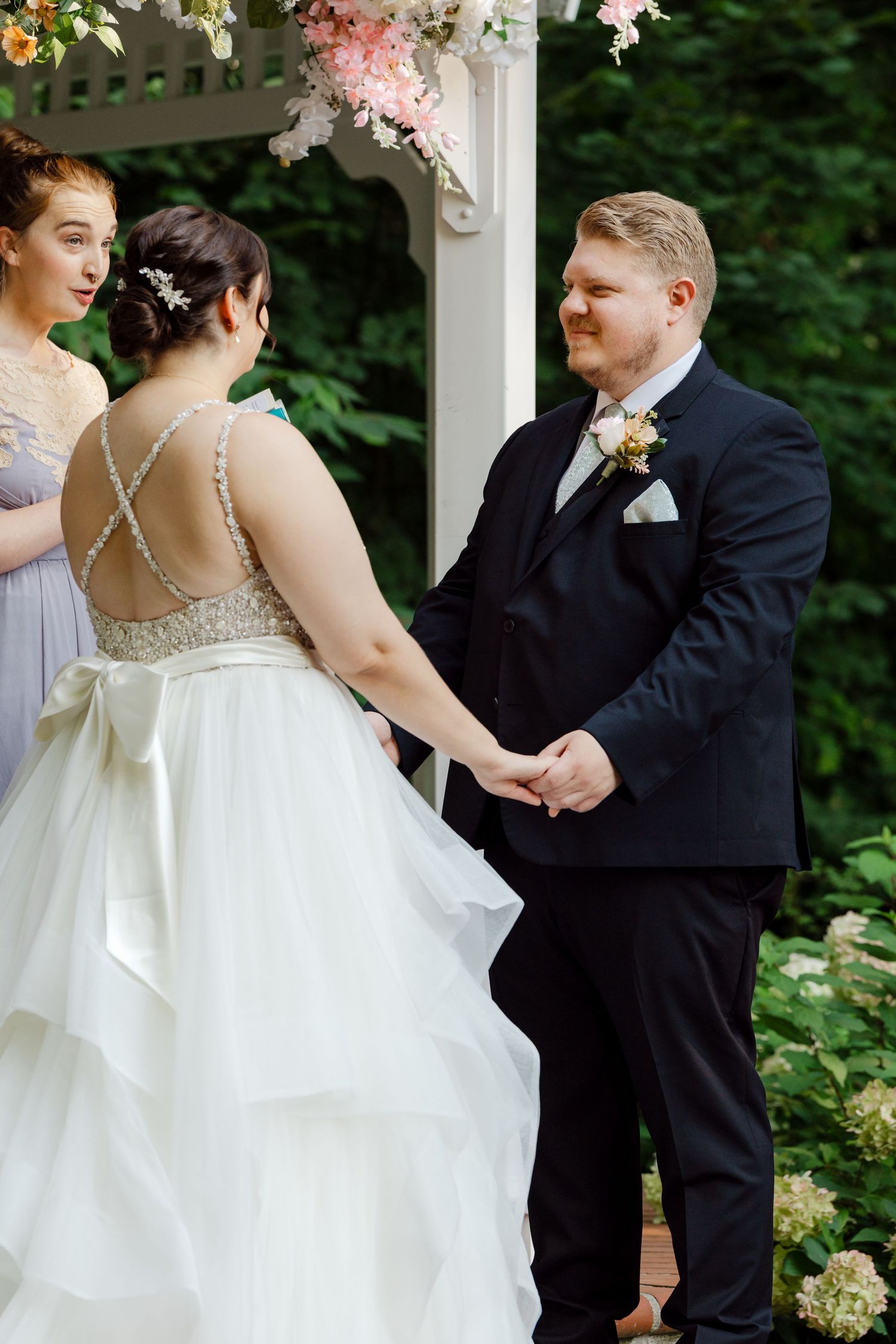 Bride in beaded backless gown holds hands with groom in navy suit at outdoor gazebo wedding ceremony.