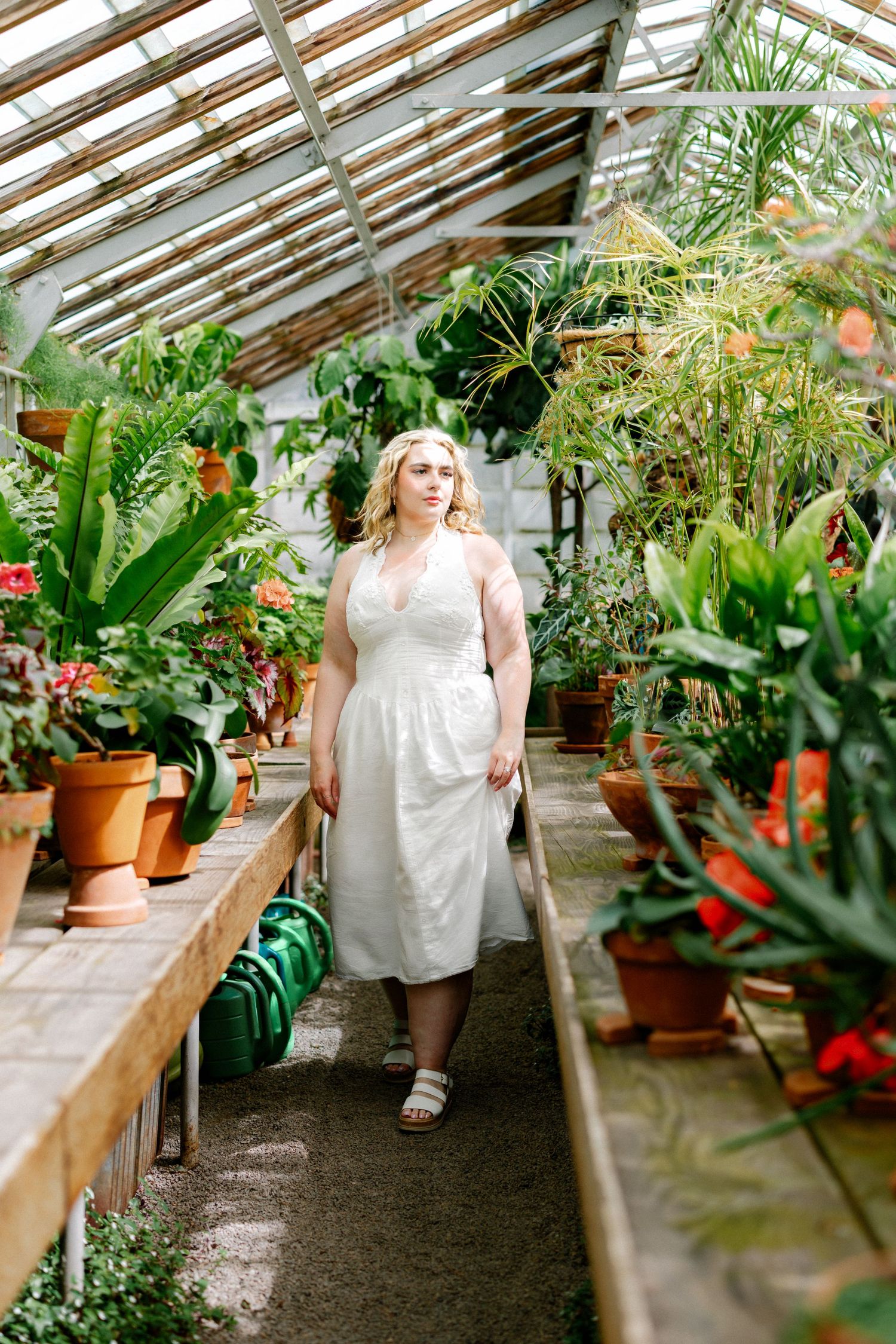 Woman in white dress walking through a lush greenhouse surrounded by potted plants and tropical foliage.
