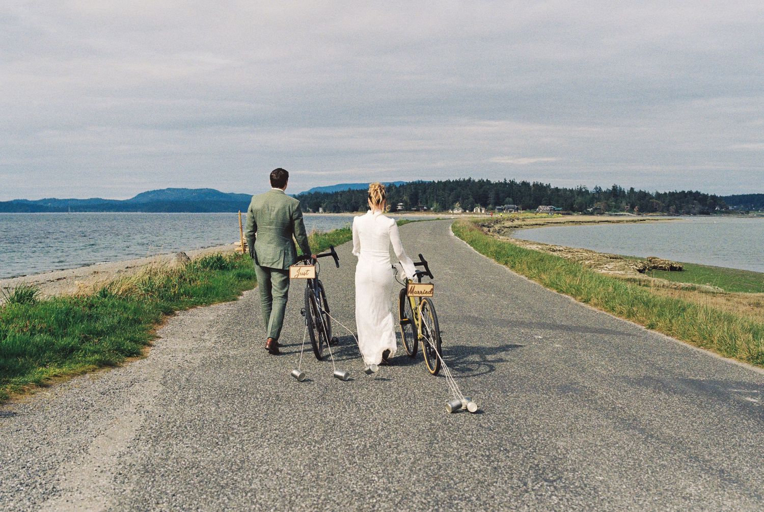 Bride and groom walk bicycles along scenic coastal road with ocean and islands visible in background.