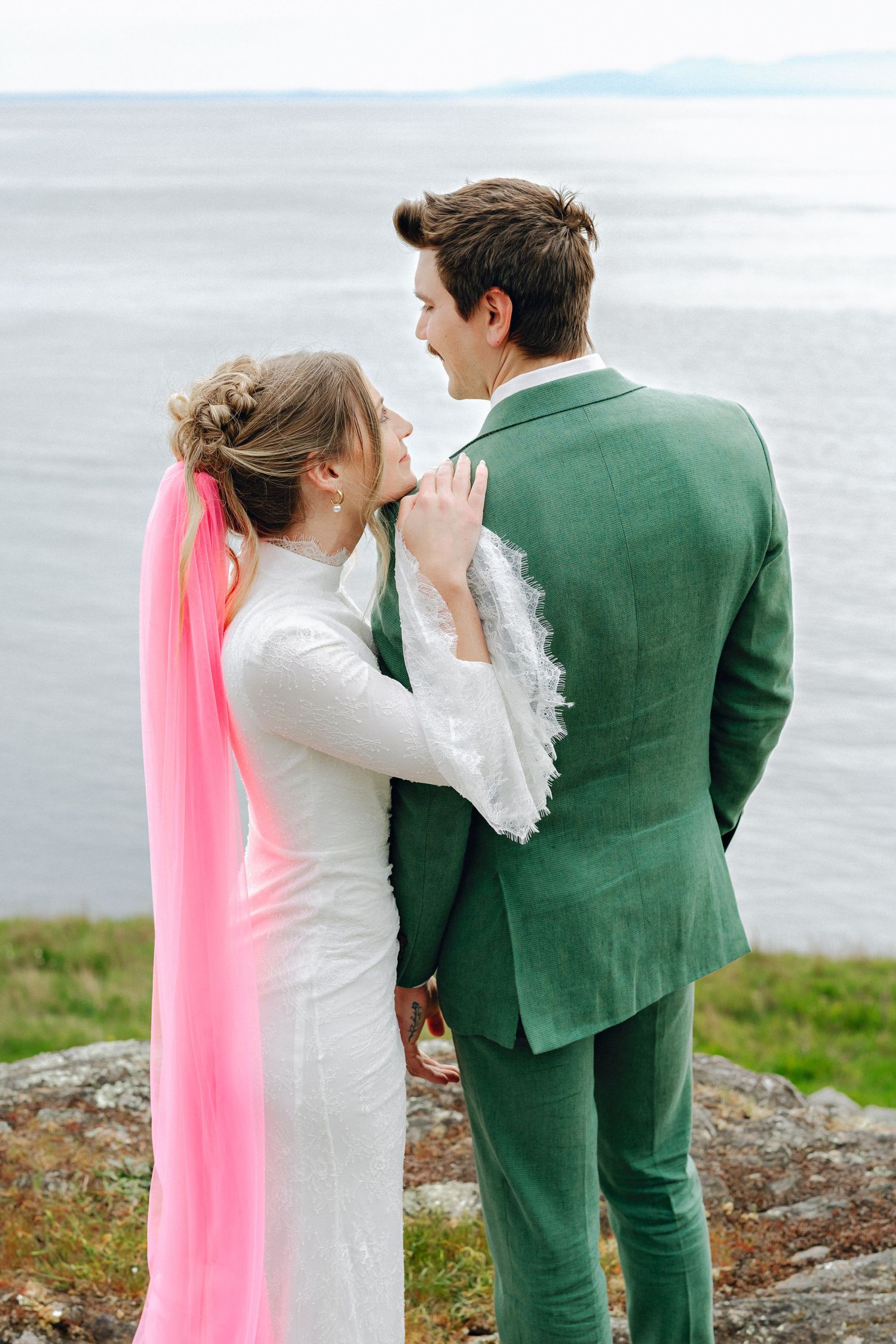 Bride in white dress with pink veil and groom in green suit stand together overlooking the ocean.