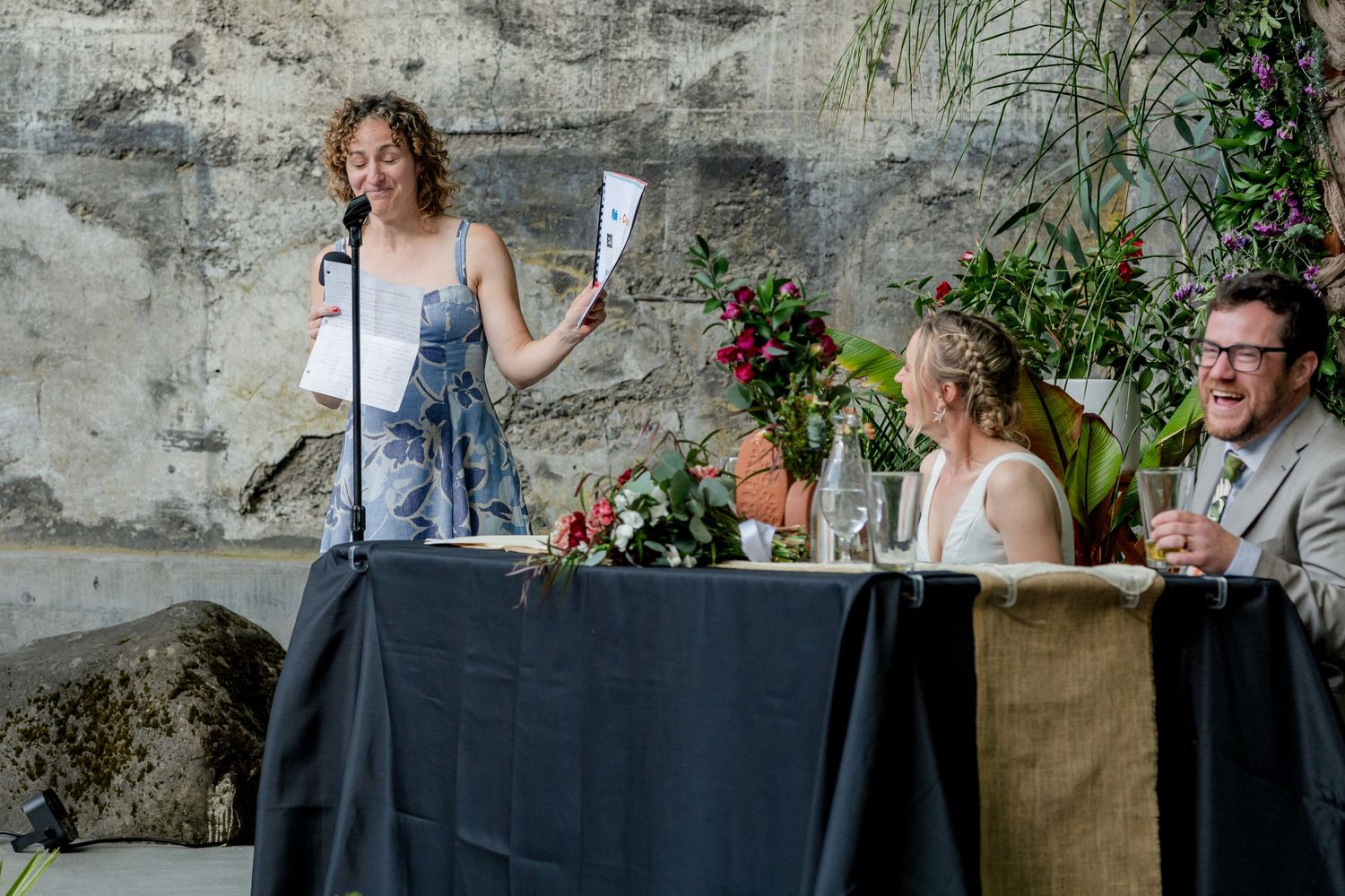 A woman reads a speech at a wedding reception while the bride and groom smile at the head table.