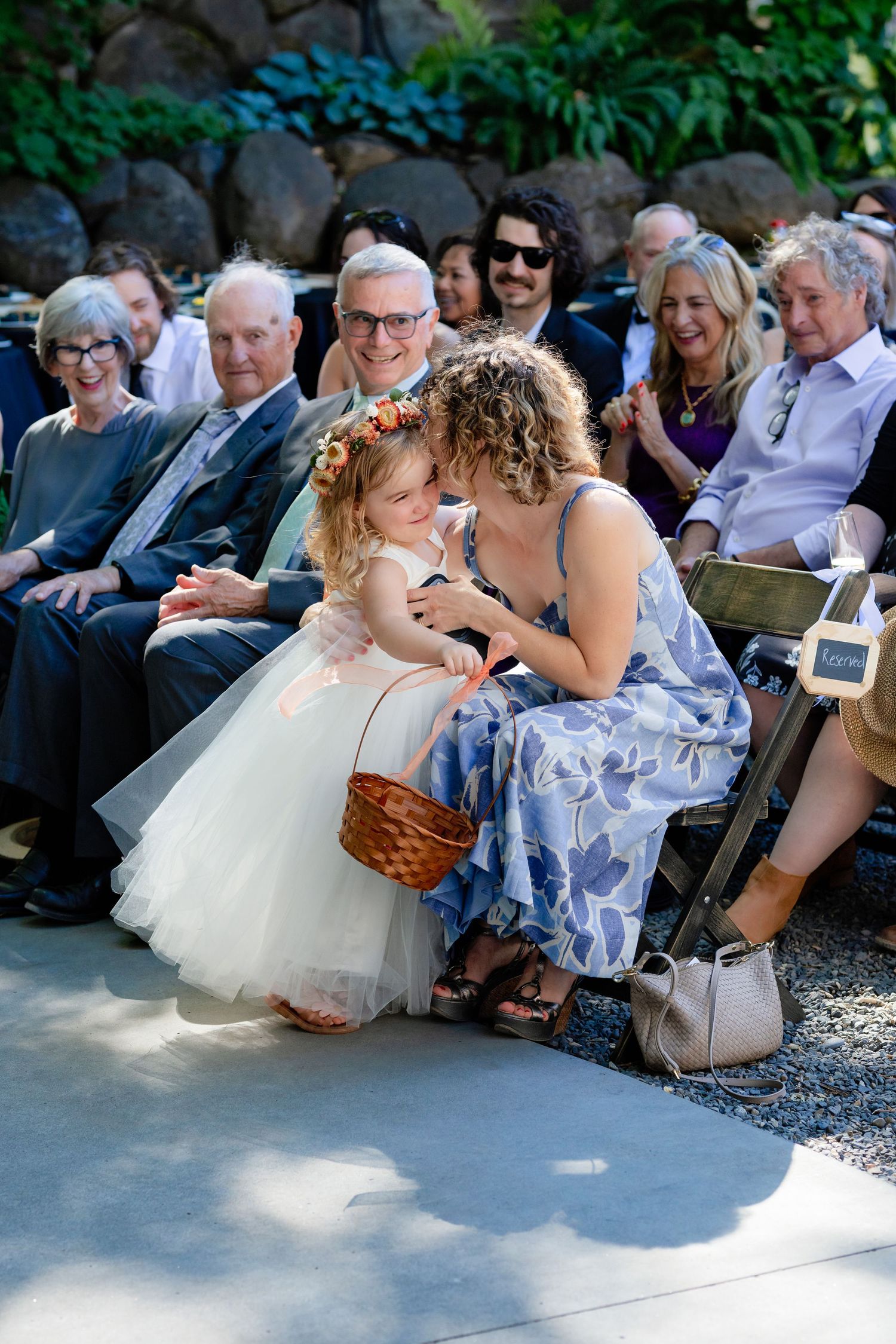 Flower girl in white tutu dress with floral crown receives kiss from seated wedding guest.