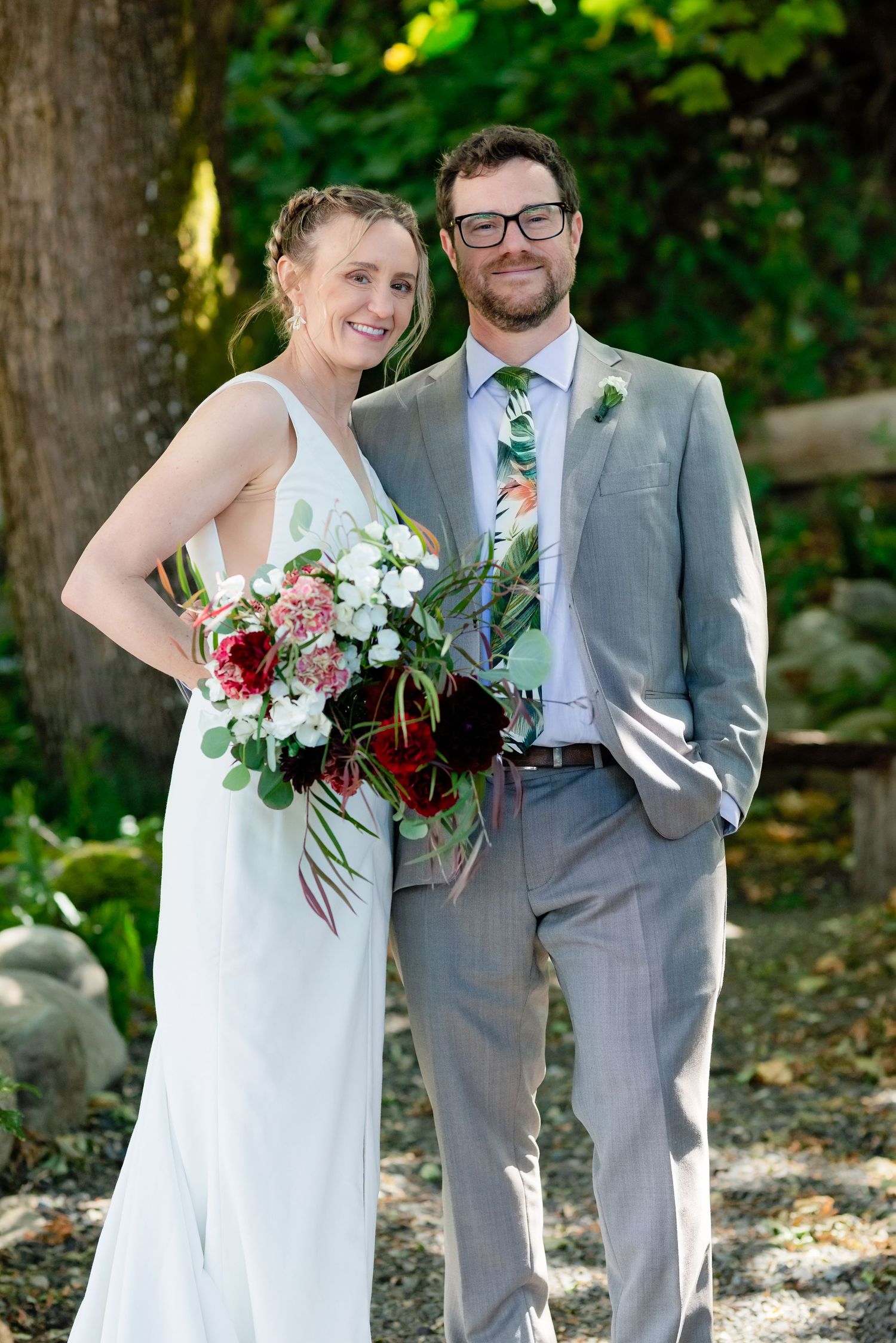 Smiling bride and groom pose outdoors, she holds a lush bouquet of red and white flowers, he wears a gray suit with floral tie.