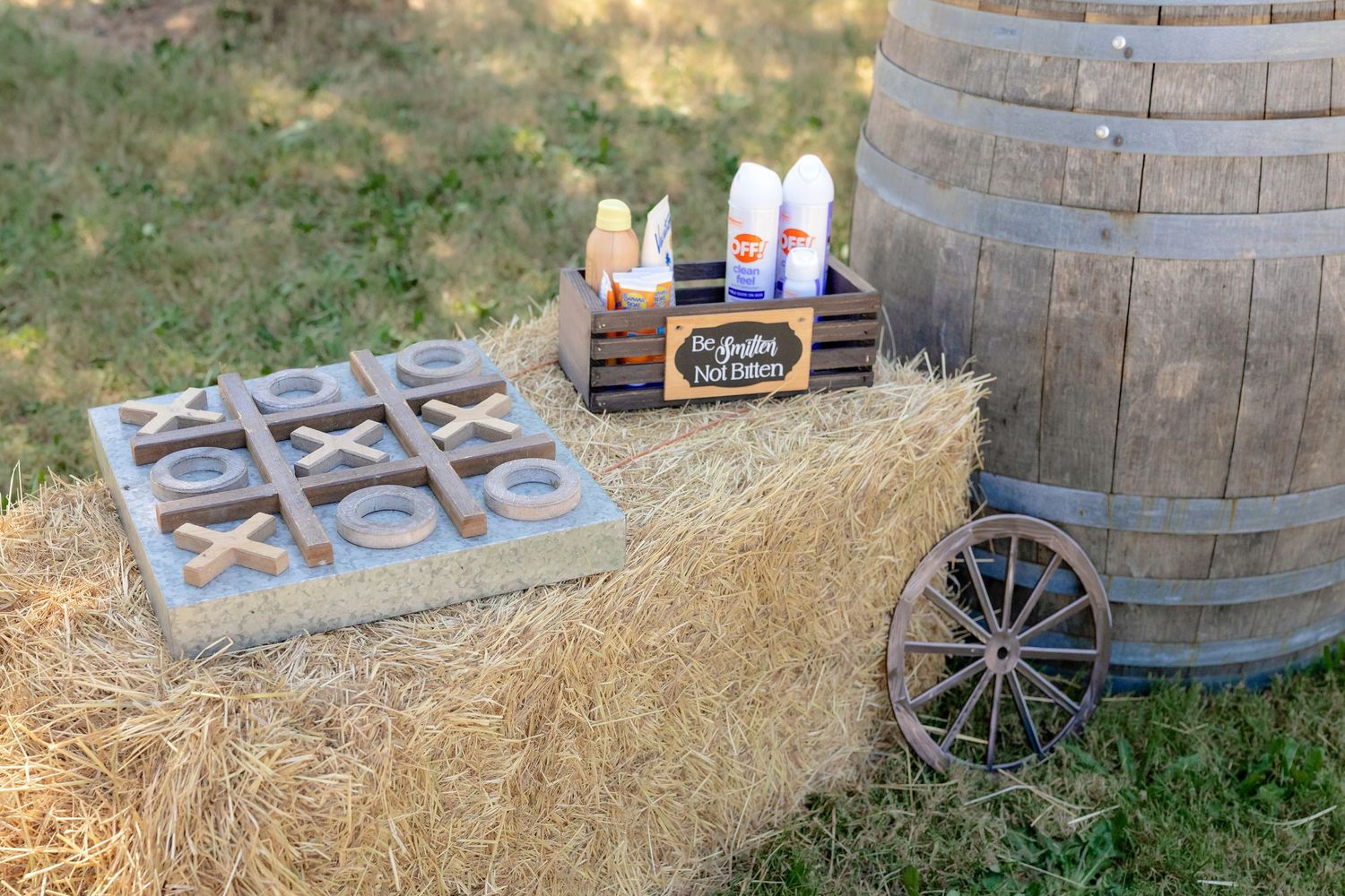 Rustic outdoor wedding decor with hay bale, oversized tic-tac-toe game, wooden crate, and barrel.