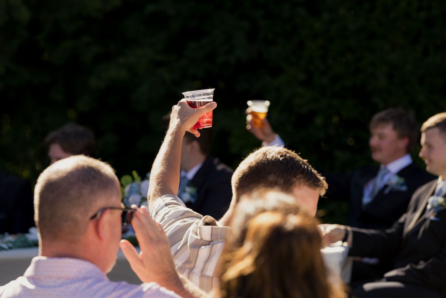 Wedding guests raise glasses in a toast outdoors in bright sunlight during reception.