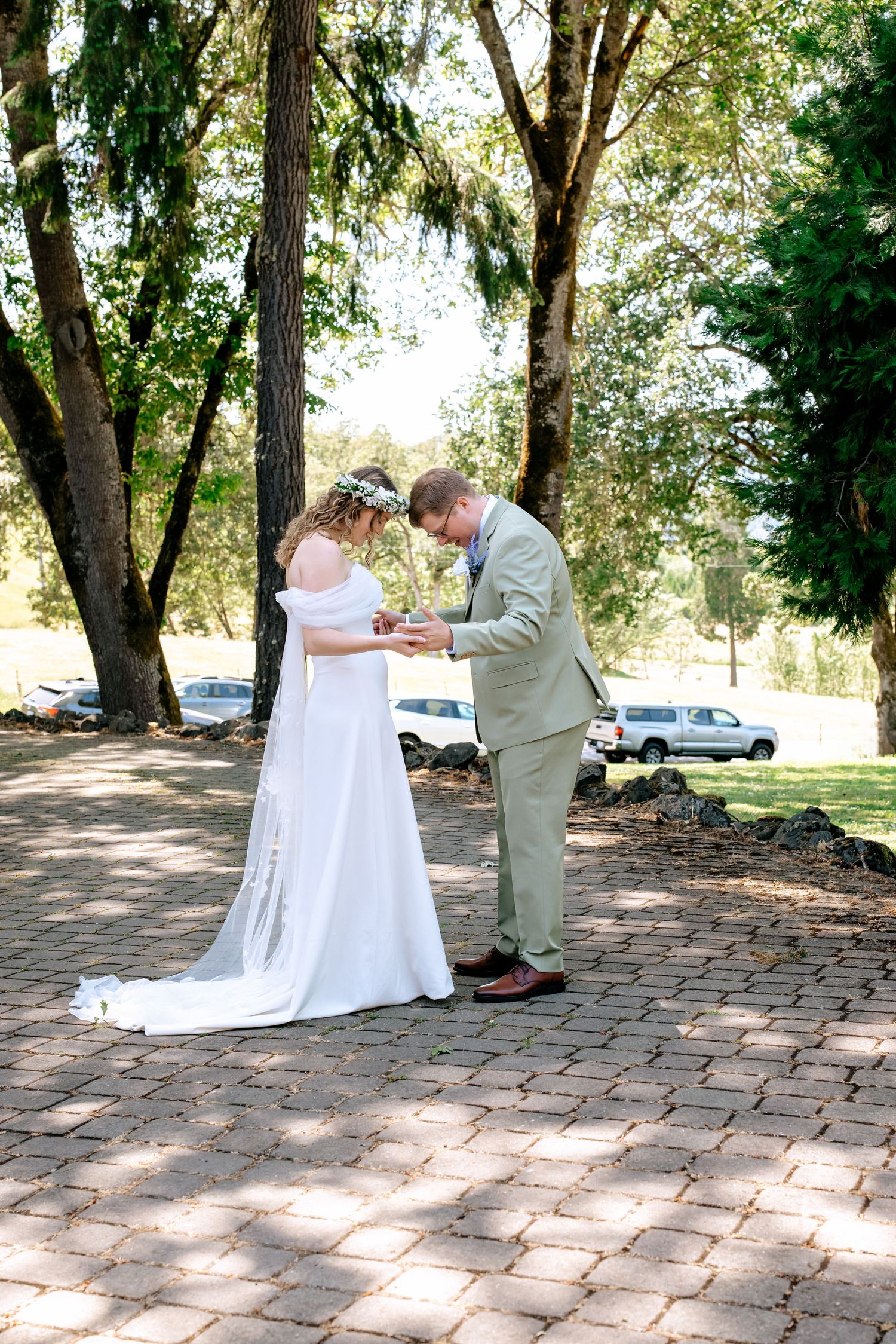 Bride and groom share an intimate first look moment holding hands on brick patio surrounded by tall trees.
