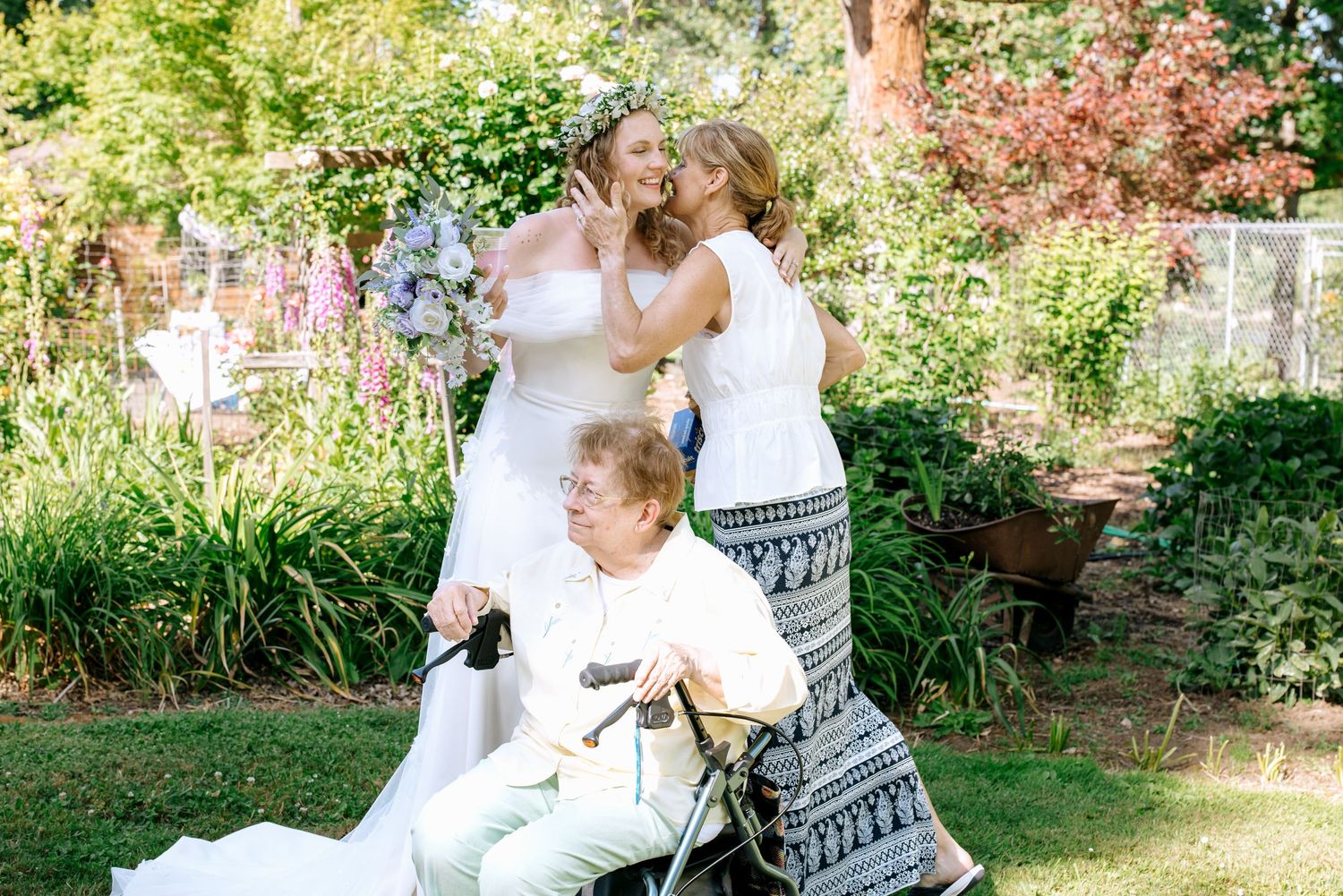 Bride shares a joyful moment with two women, one seated in a wheelchair, in a lush garden.