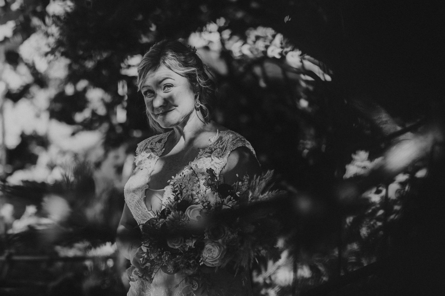 Black and white bridal portrait of a smiling woman holding a bouquet, surrounded by lush garden foliage.