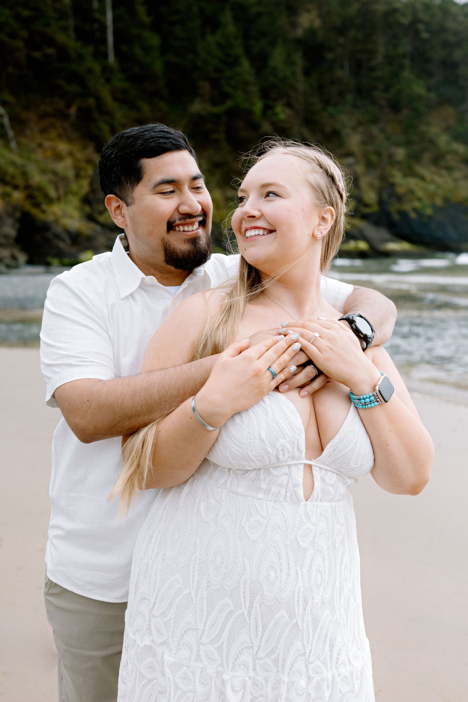 Man embracing woman in white dress on a beach, both smiling joyfully with ocean and forest backdrop.