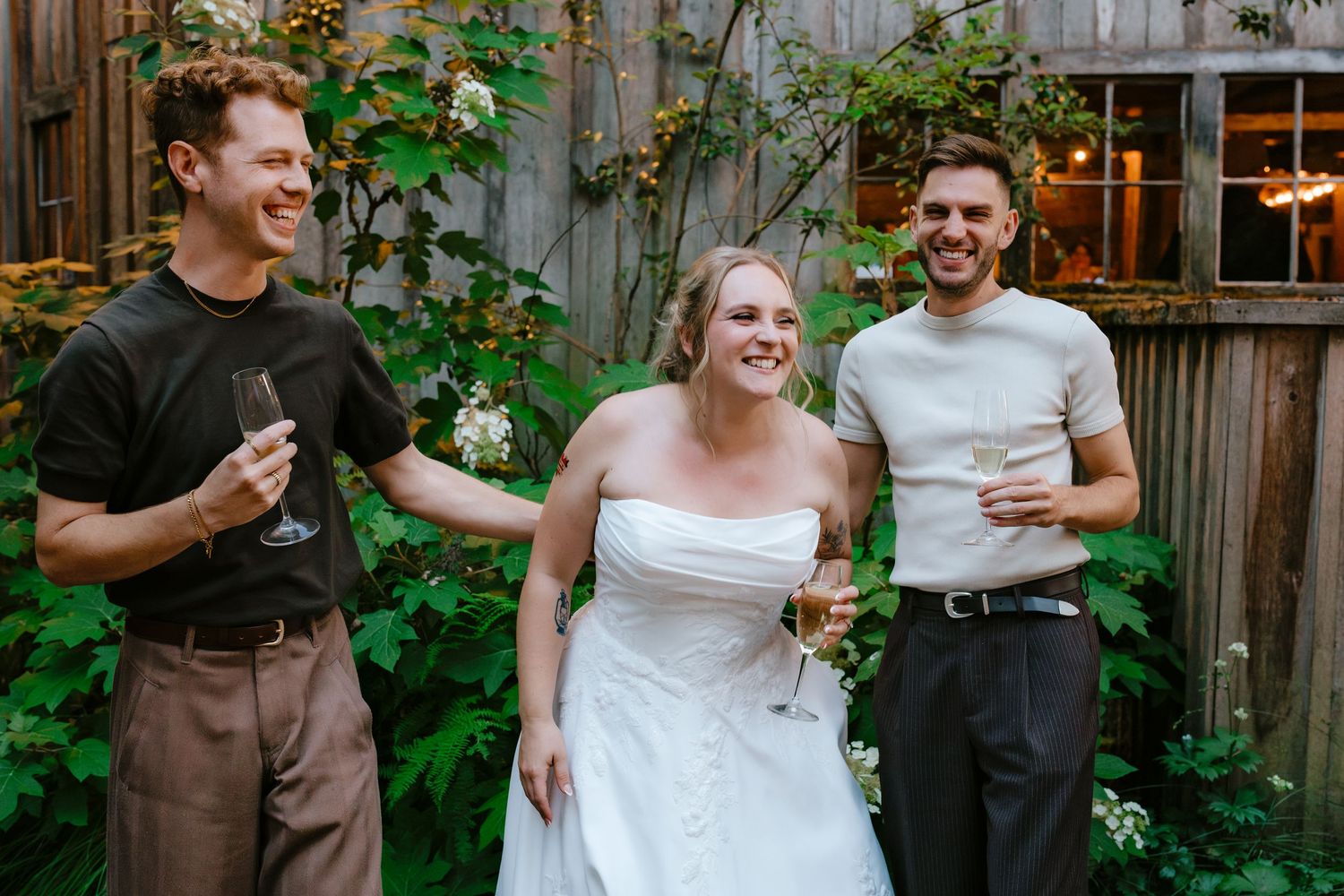Bride in white strapless gown laughing with two male guests holding wine glasses outdoors.
