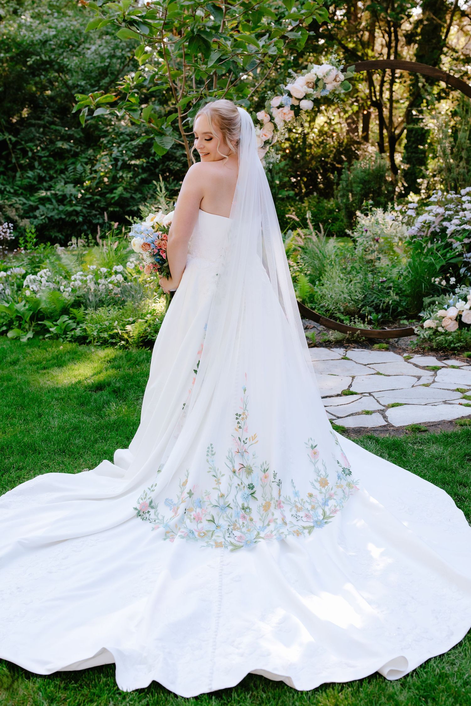 Bride in floral embroidered wedding gown with cathedral veil stands in lush garden holding bouquet.