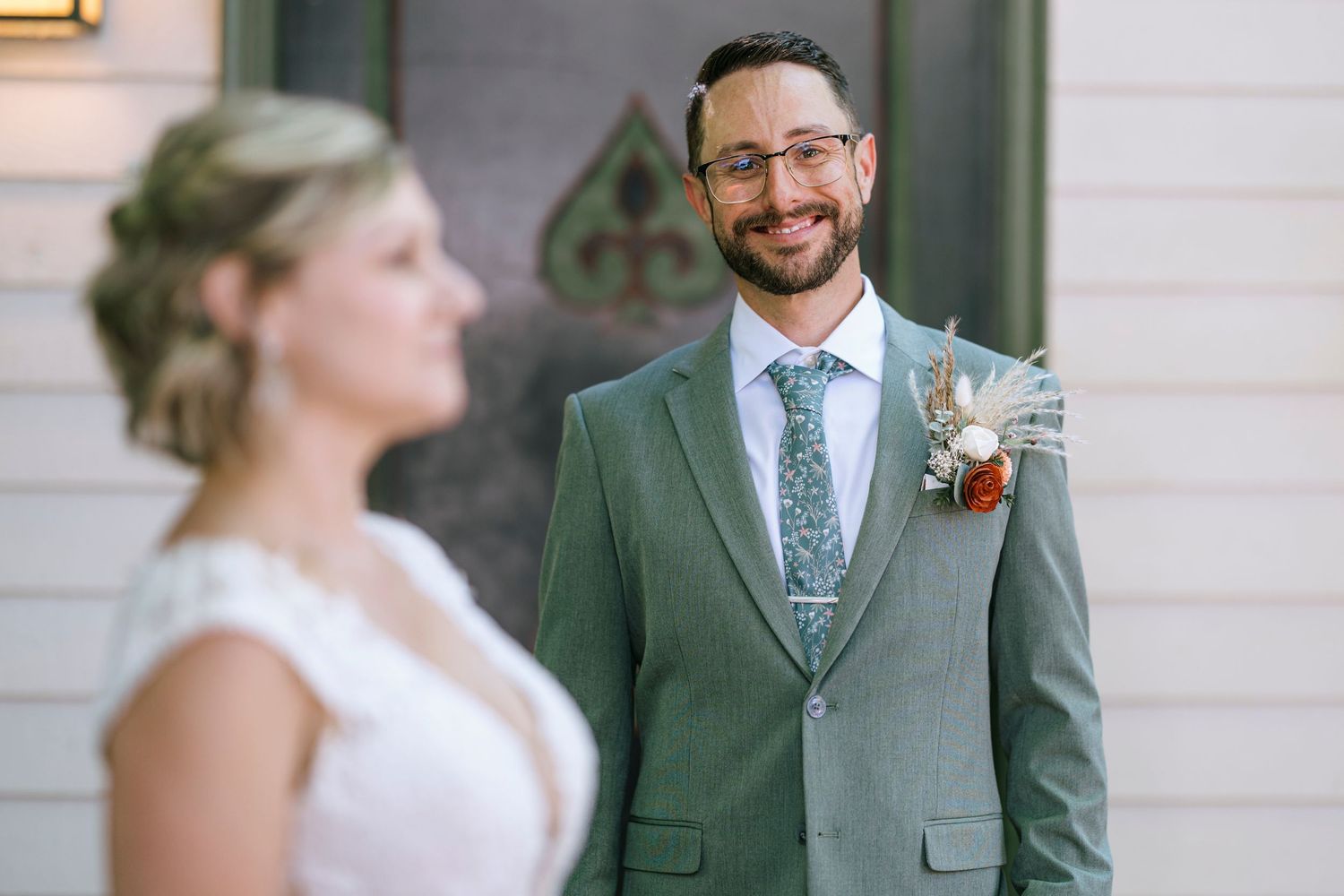 A wedding couple in elegant attire poses outdoors, with a sage green suit and white dress creating a classic look.