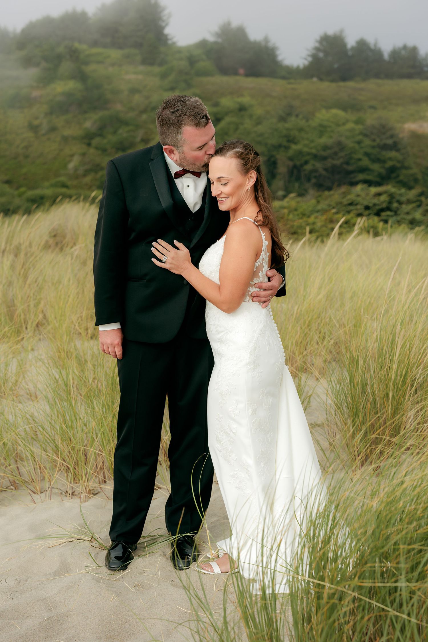 Wedding couple embracing in tall beach grass on a misty coastal hillside.