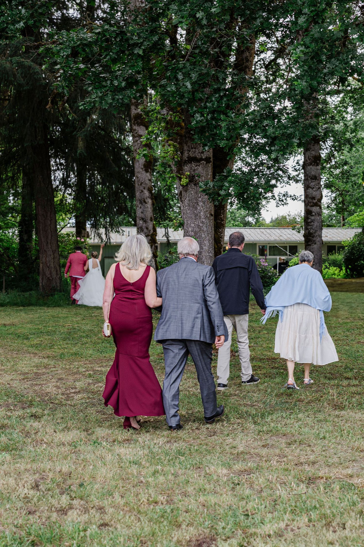 Wedding guests in formal attire walk across a grassy lawn surrounded by tall trees.