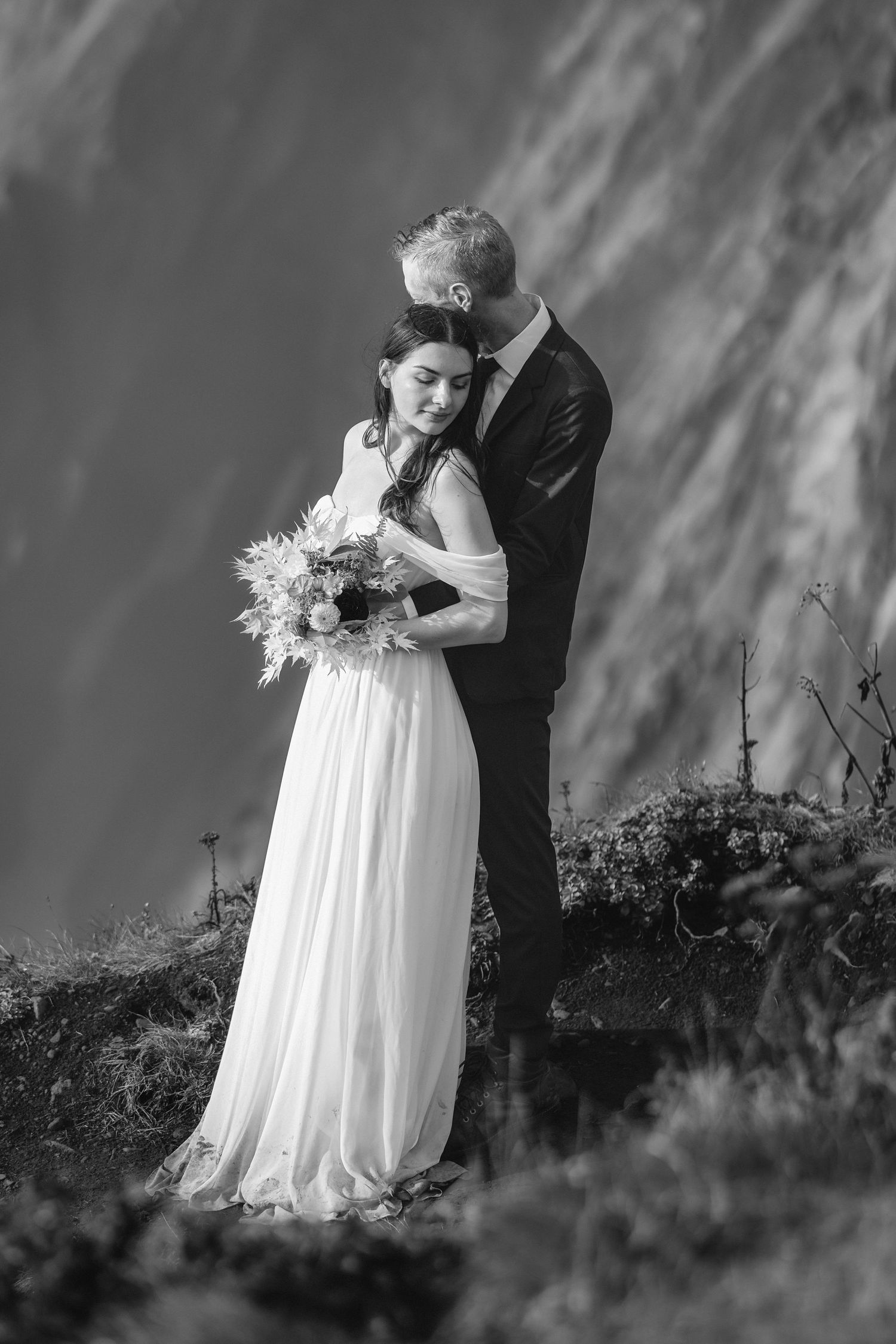 A romantic black and white wedding portrait captured against a dramatic mountainside backdrop.
