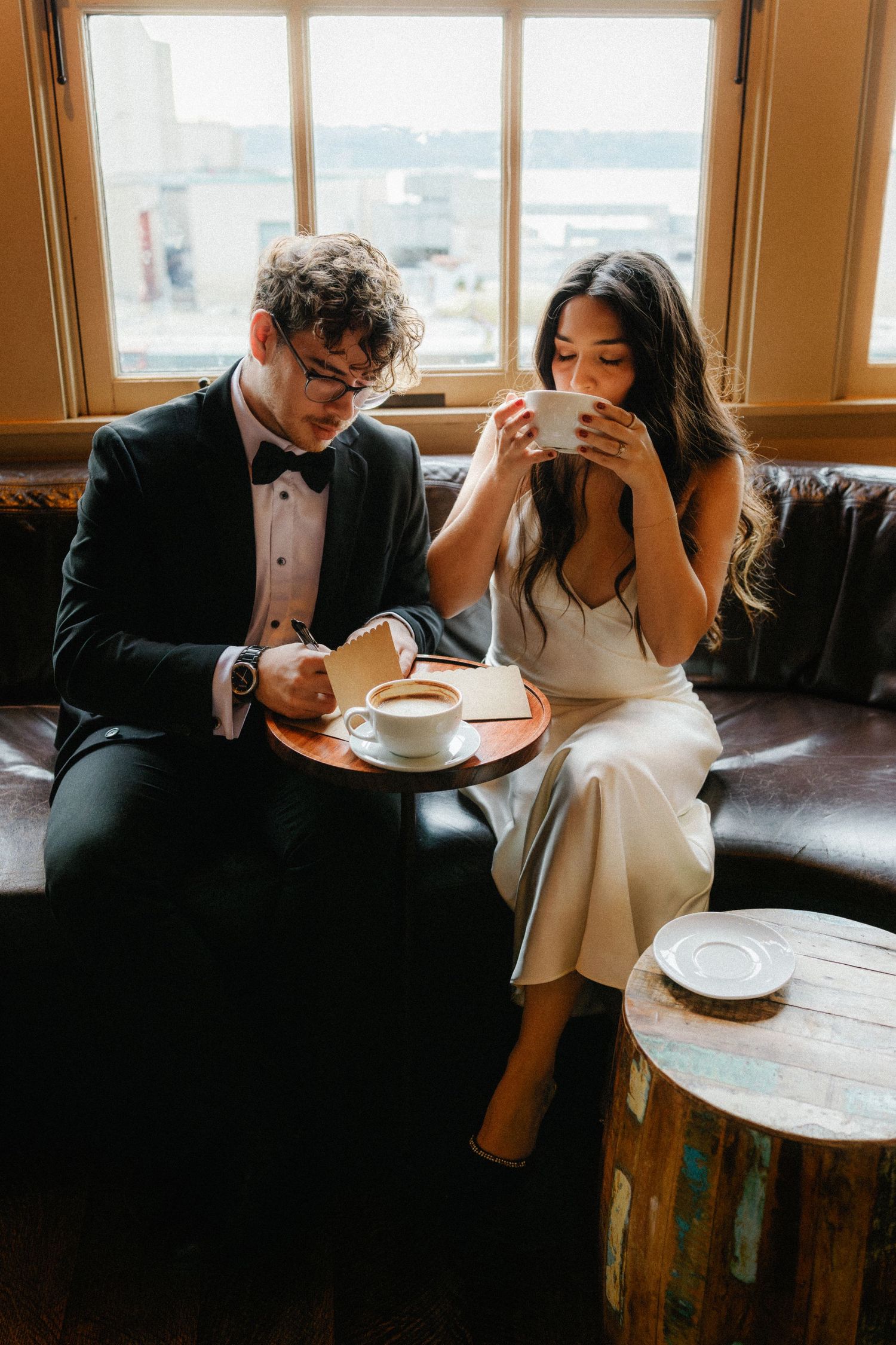 A couple enjoys coffee together on a leather couch by a window in formal attire.
