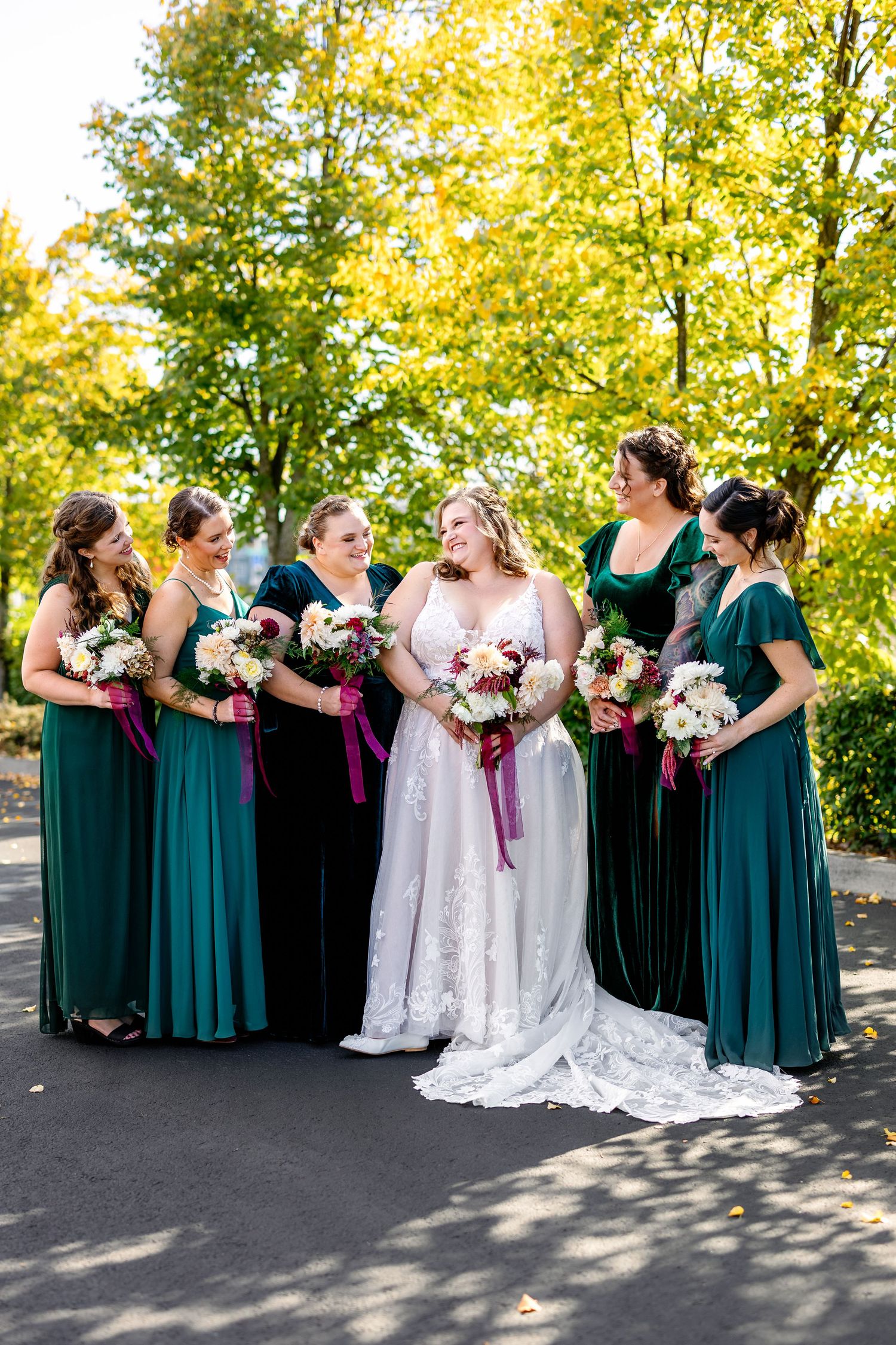 Wedding party in emerald green dresses and white gown posing outdoors under autumn trees.