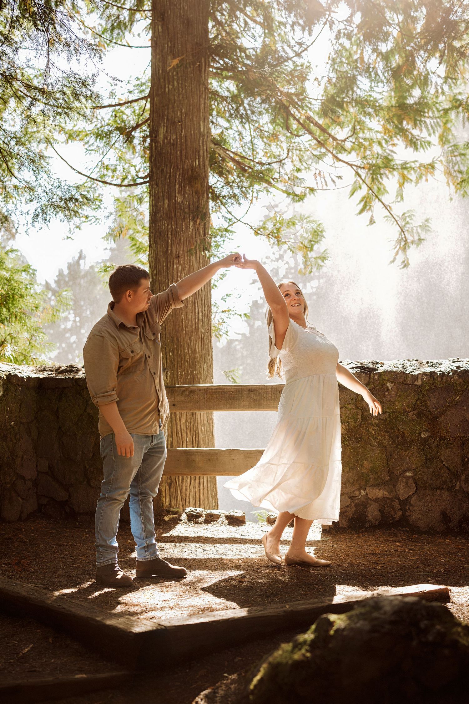 A couple dances playfully in sunlit forest clearing beside a stone wall.
