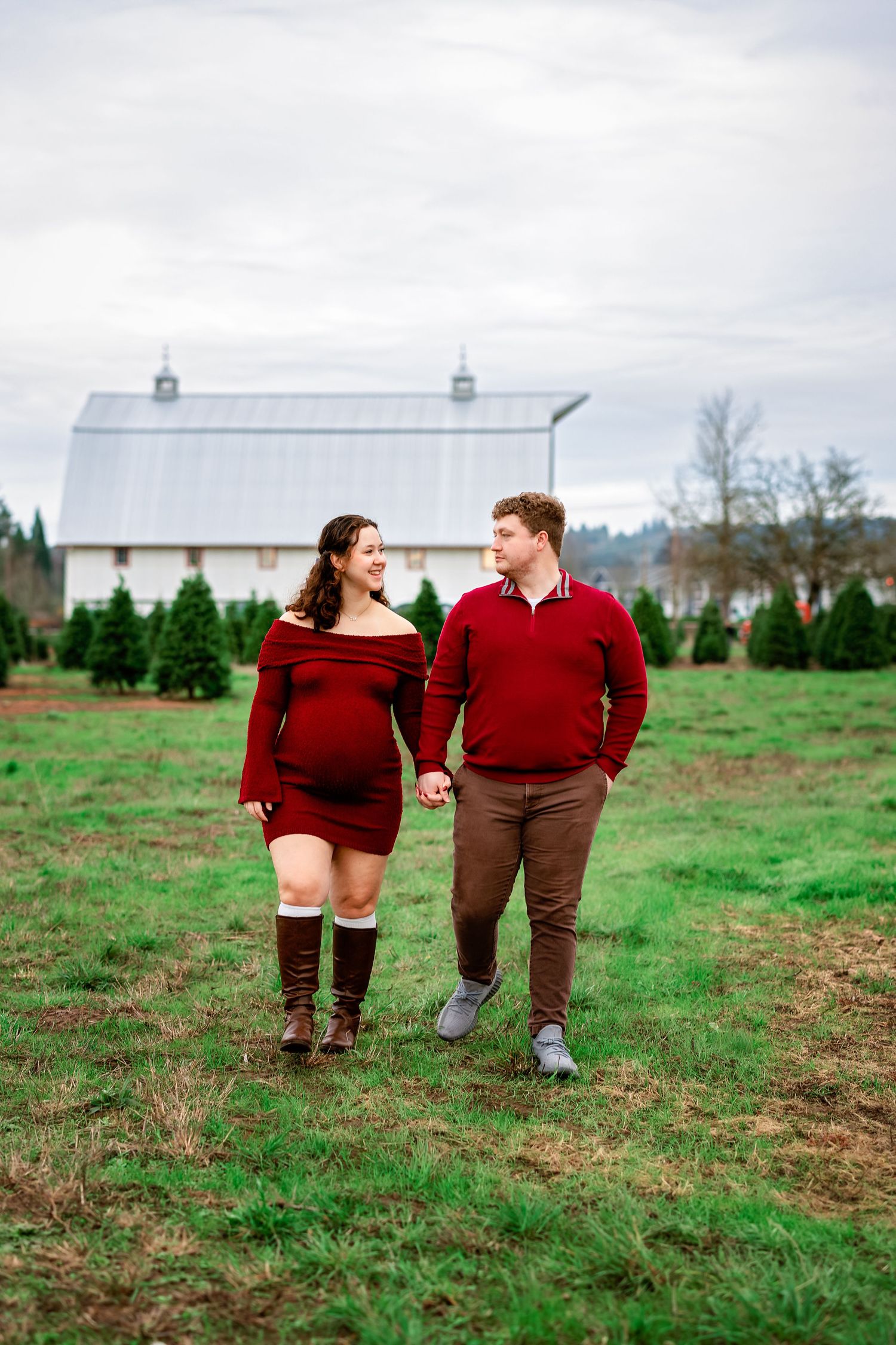 A couple wearing burgundy sweaters walks together in a green field with a white barn in the background.
