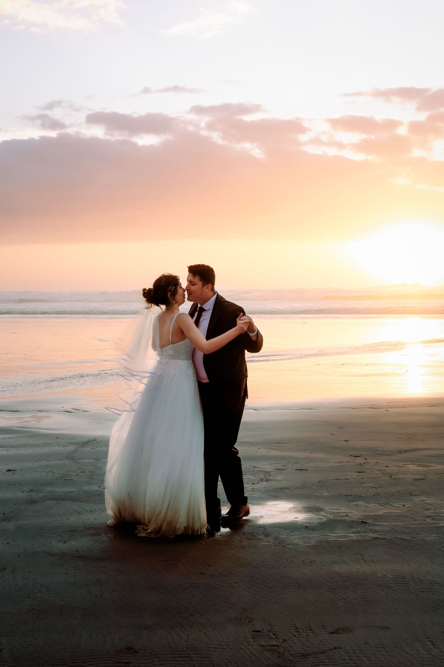 A wedding couple dances on the beach at sunset with waves gently rolling in the background.