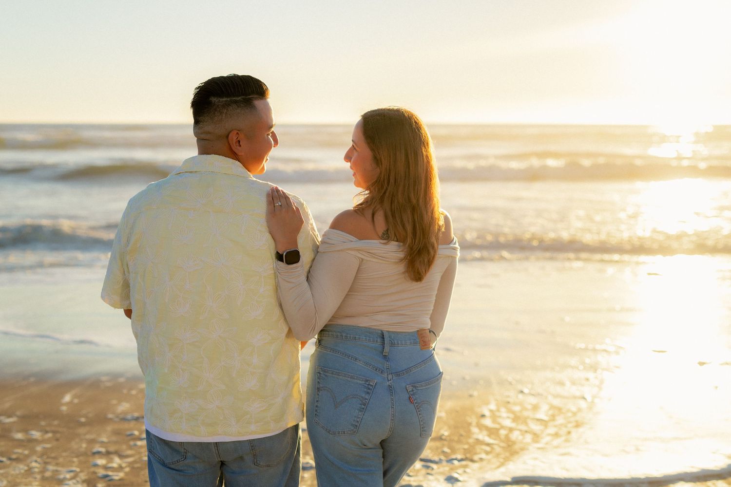 Couple standing together on a beach at sunset, facing each other with ocean waves in the background.