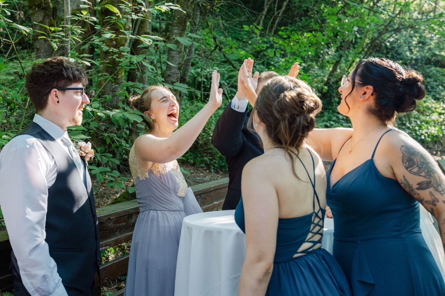 Bridesmaids and groomsman celebrate with high fives in blue and gray attire at outdoor wedding.