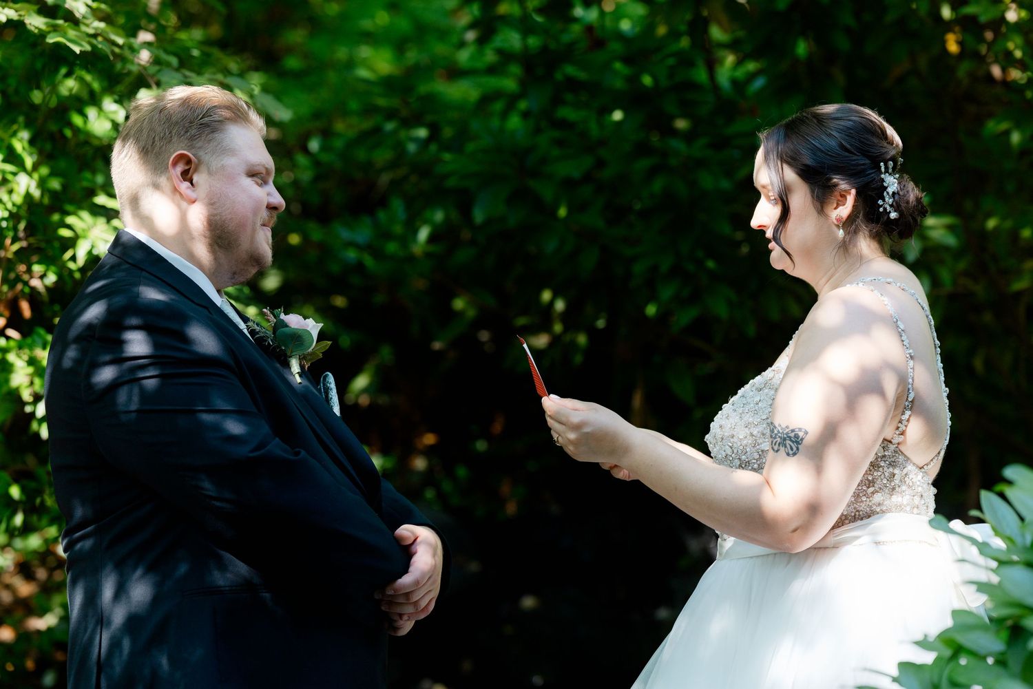 Bride reads wedding vows to groom outdoors surrounded by lush green trees during intimate ceremony.