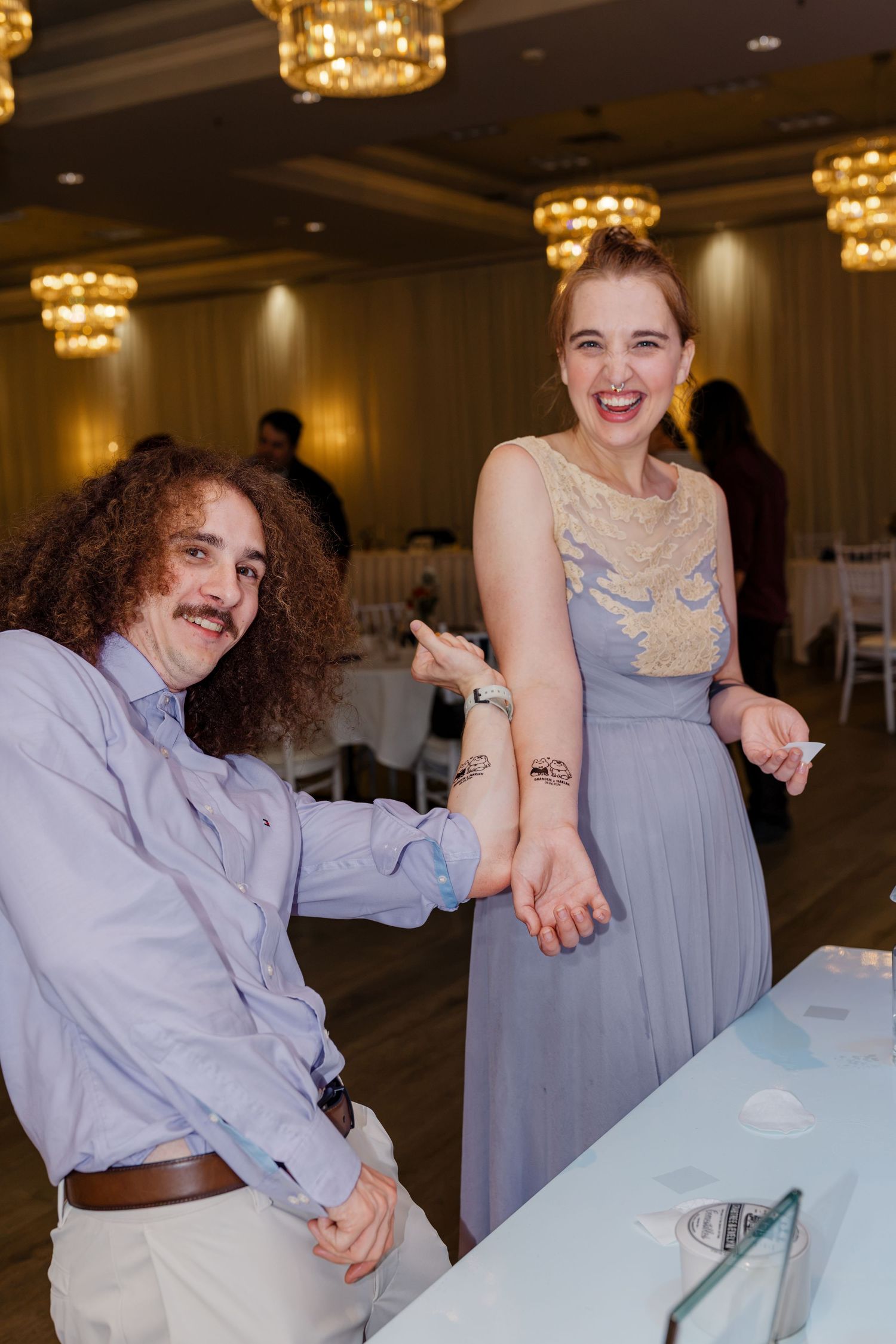 Laughing couple shows off matching temporary tattoos at elegant wedding reception with golden chandelier lighting.