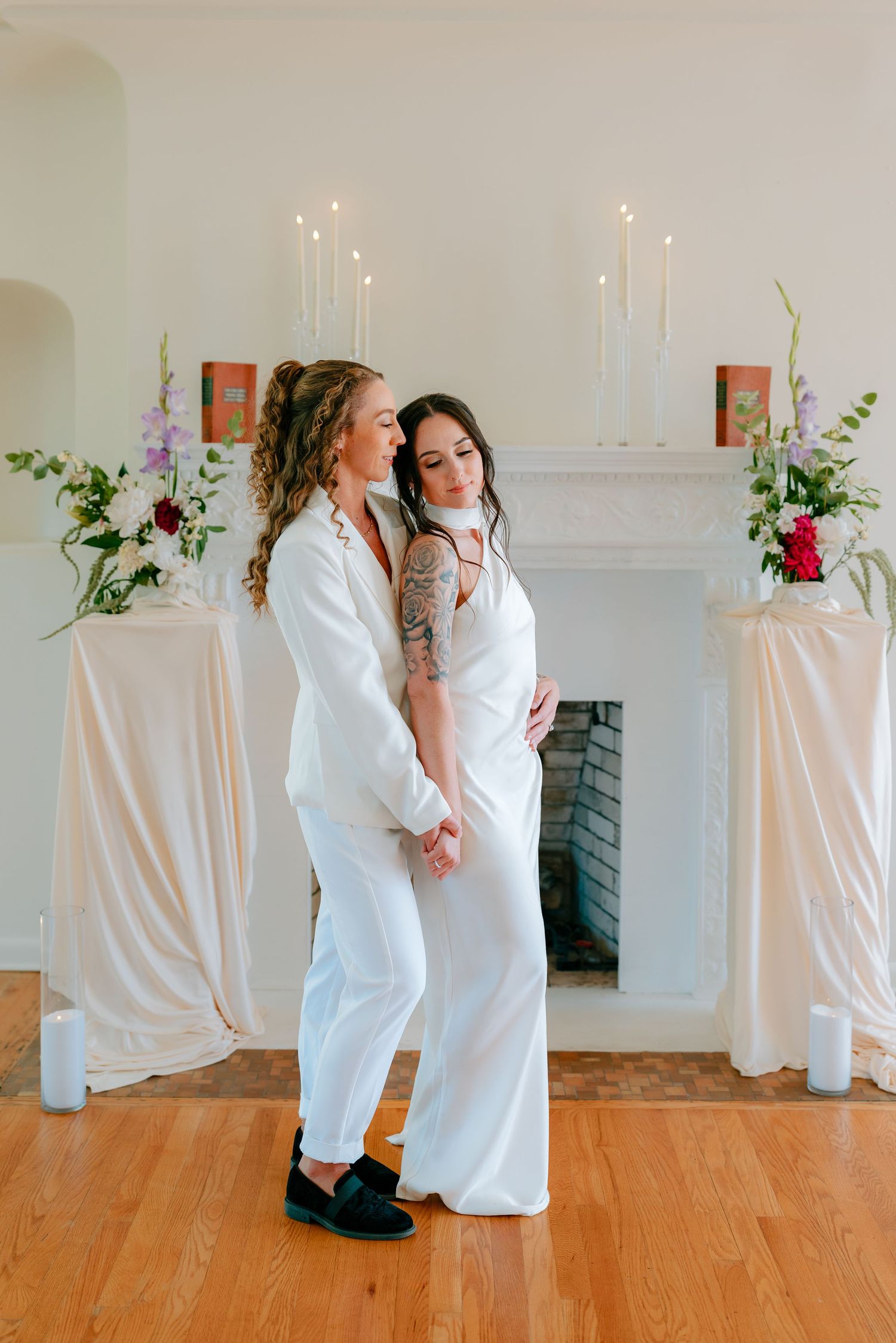 Two brides in white outfits pose together near a fireplace adorned with floral arrangements and candles.