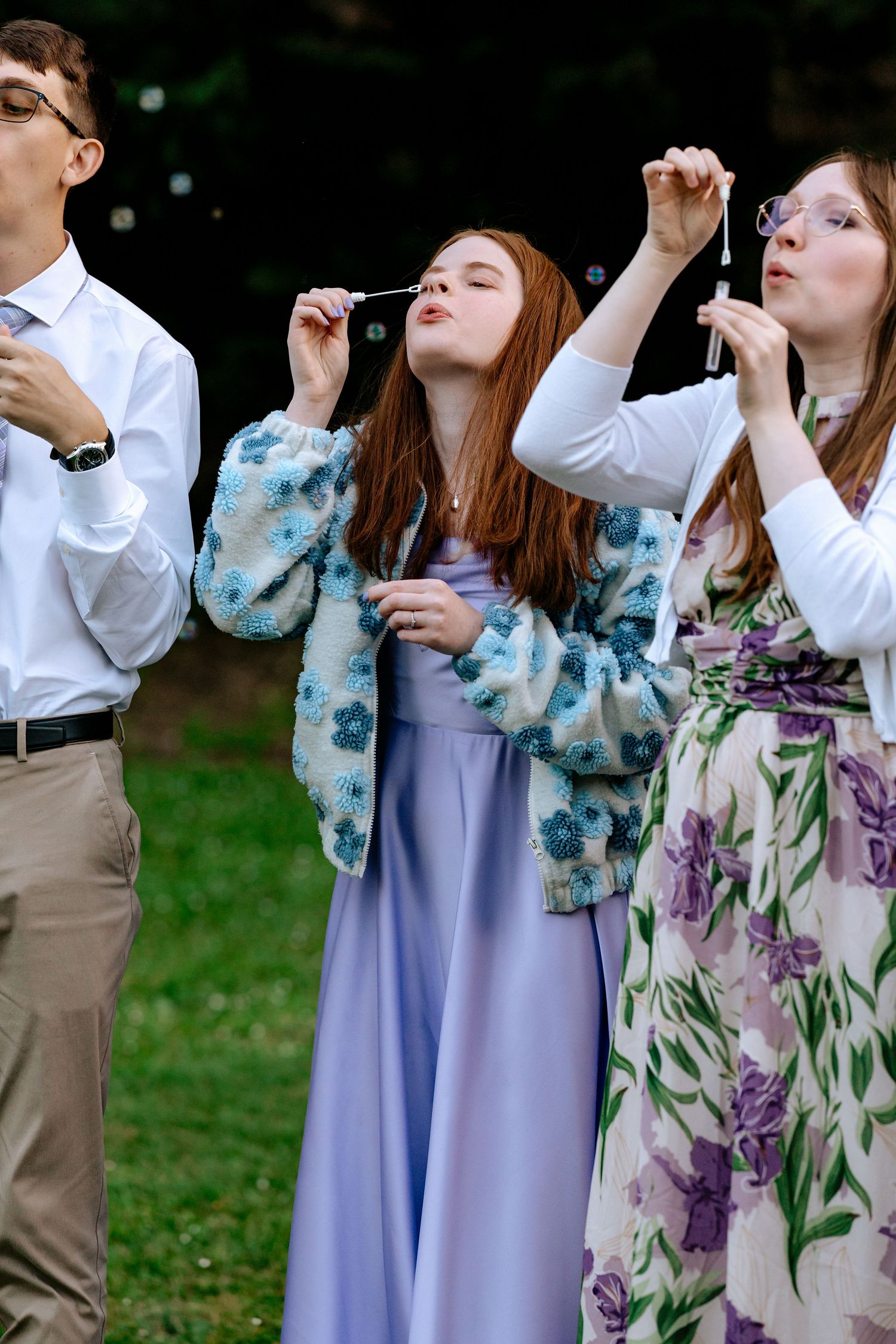 Three young wedding guests blow bubbles outdoors wearing formal attire and floral outfits.