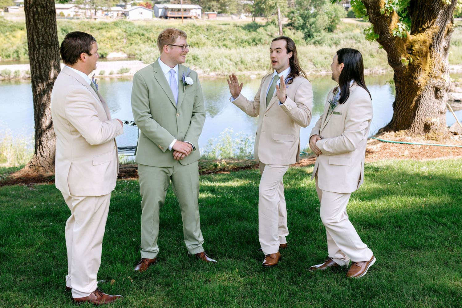 Four groomsmen in tan suits conversing outdoors near a lake on a sunny wedding day.