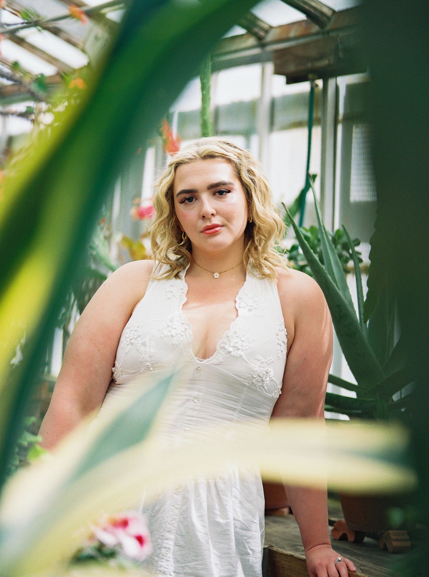 Blonde woman in white lace halter dress posing in a lush greenhouse surrounded by tropical plants.