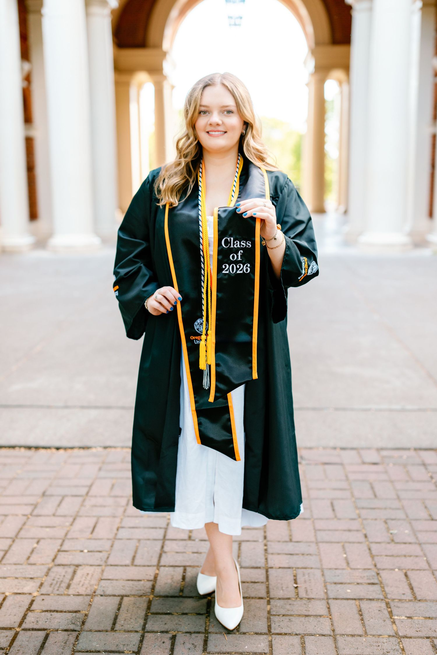 Smiling graduate in green cap and gown with gold stole reading Class of 2026 poses outdoors.