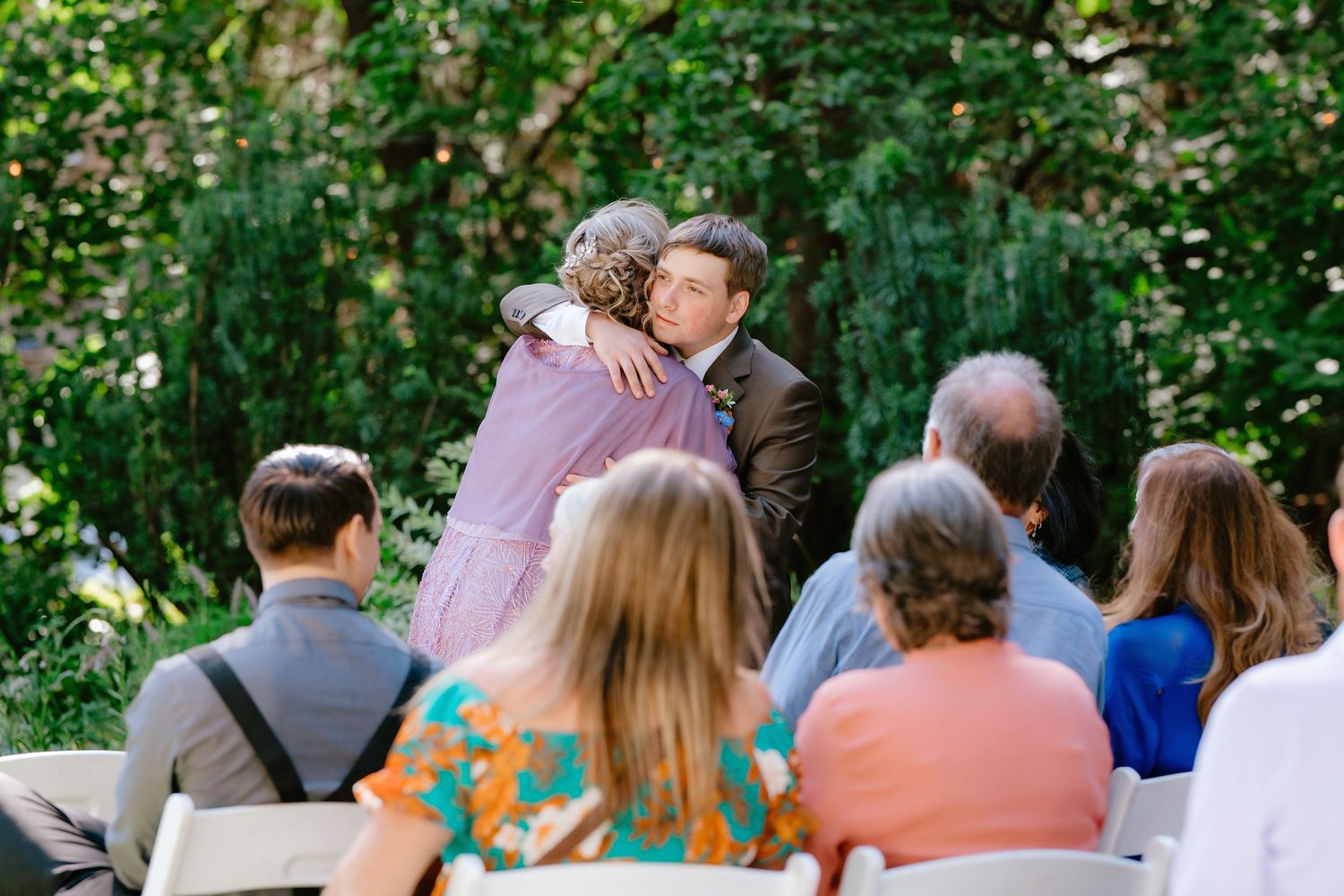 Outdoor wedding ceremony with guests seated as couple shares heartfelt hug near lush green trees.
