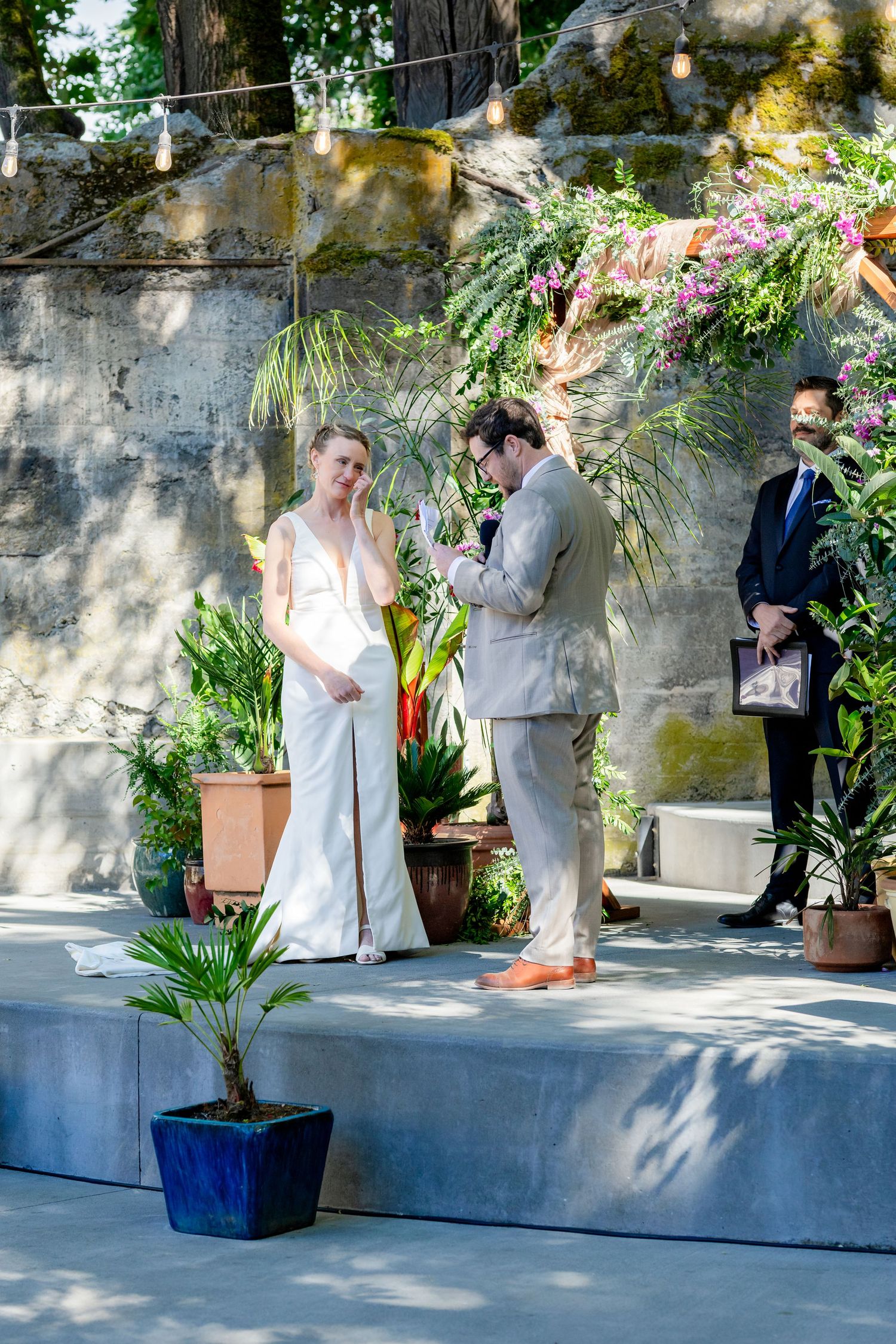 Bride and groom exchange vows outdoors at a lush garden wedding ceremony with tropical plants and string lights.