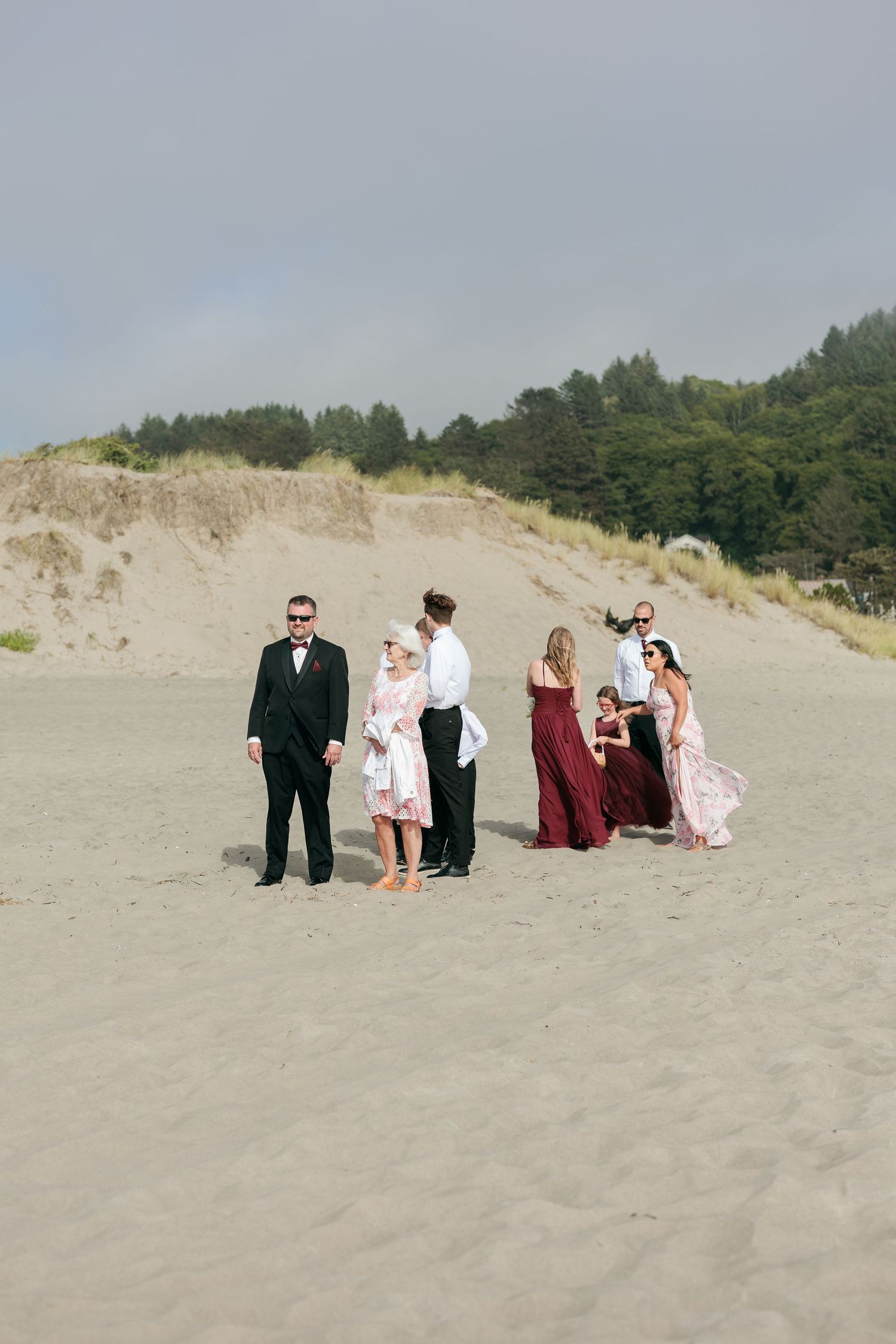 A wedding party poses on a sandy beach with forested dunes in the background.