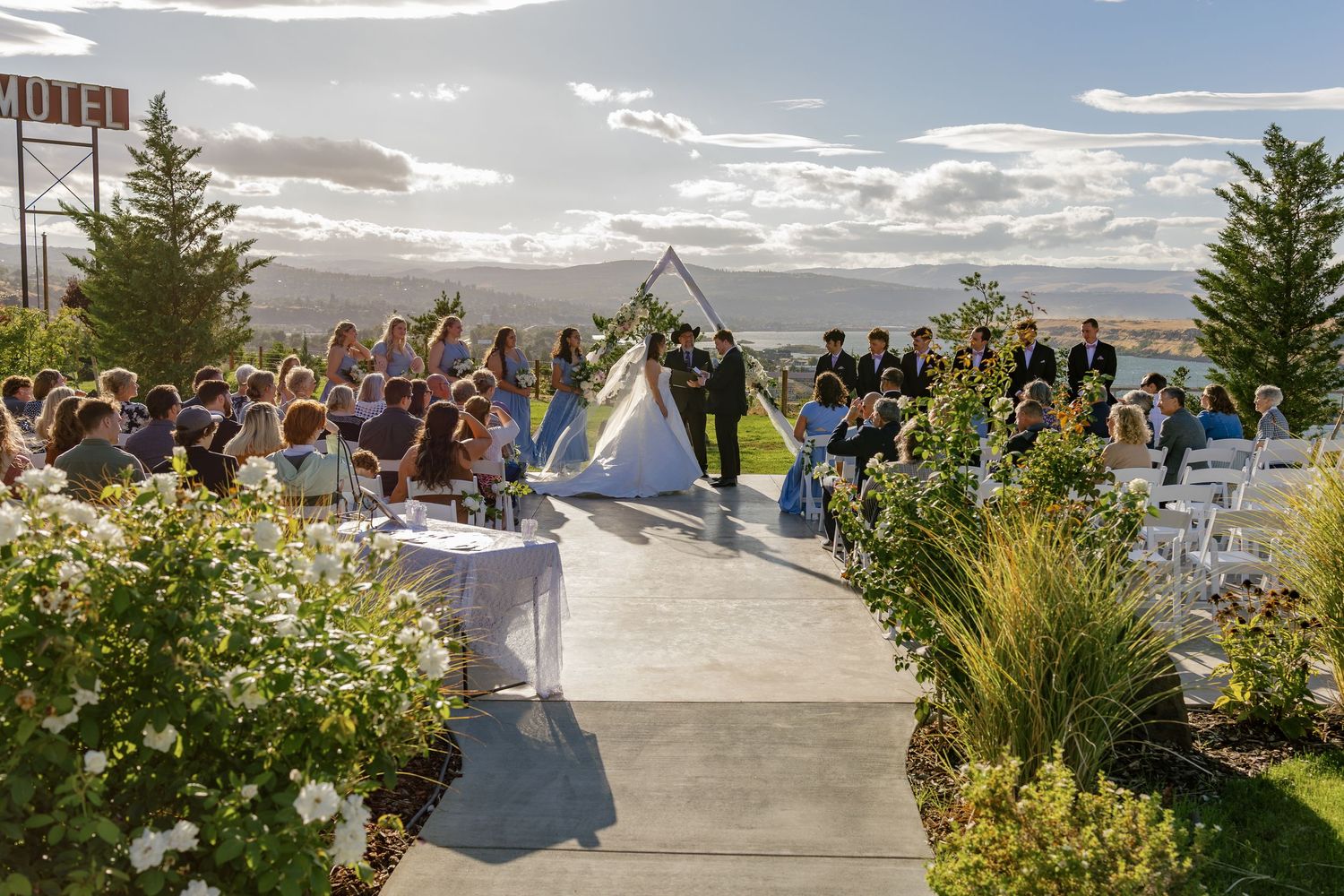 Outdoor wedding ceremony setup with mountain views and guests seated on a hillside.