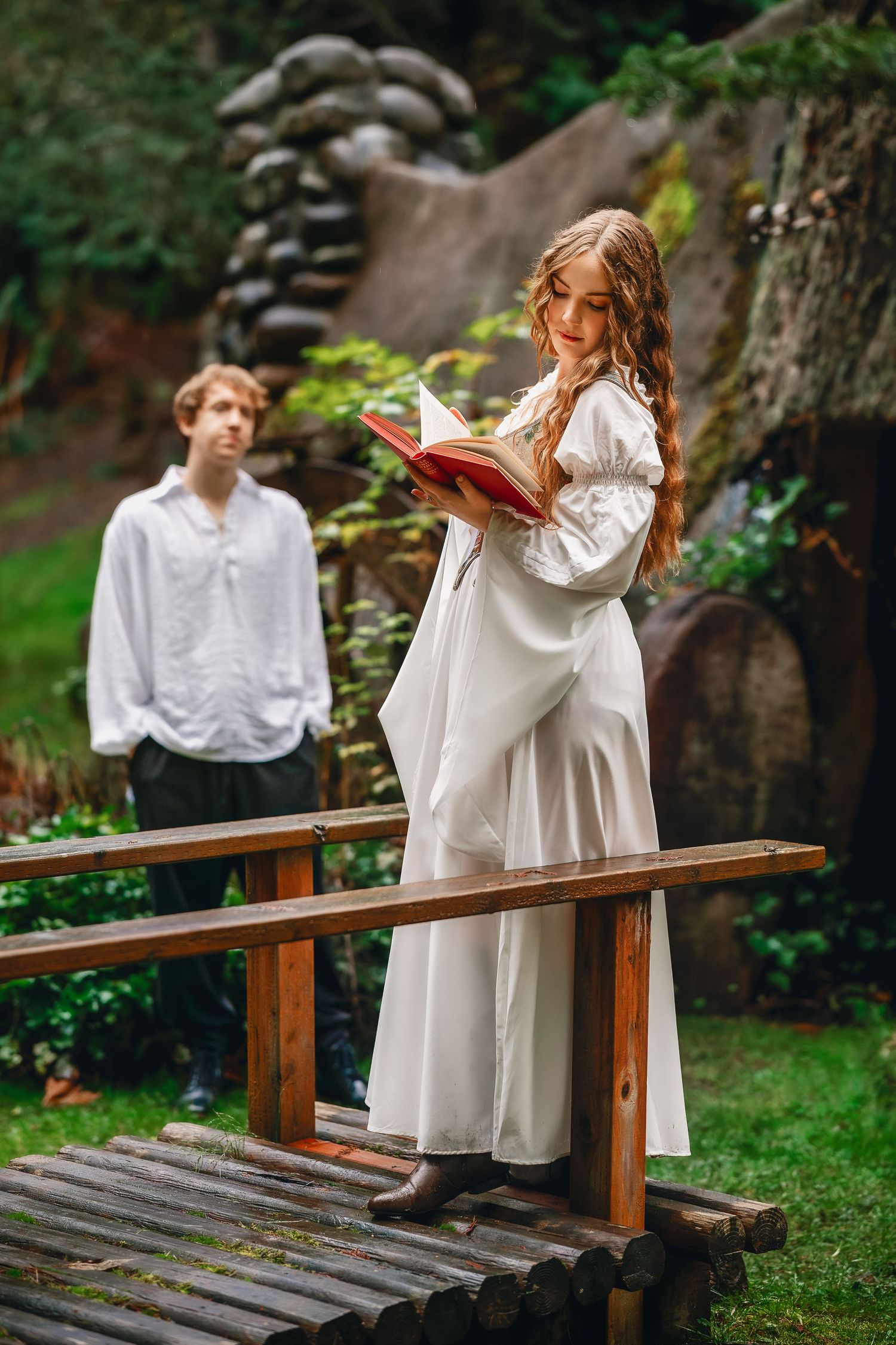 Figures in flowing white clothing sit reading on a wooden bench in a lush garden setting.