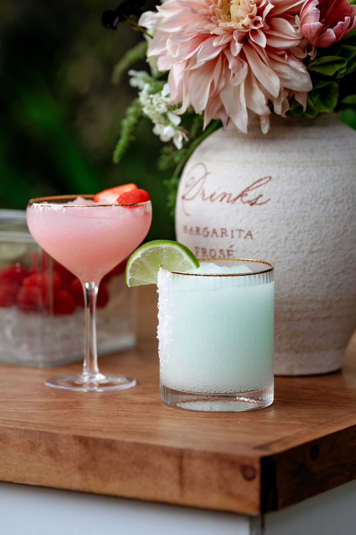 Pink and white cocktails served in elegant glassware alongside floral arrangements on a wooden serving board.