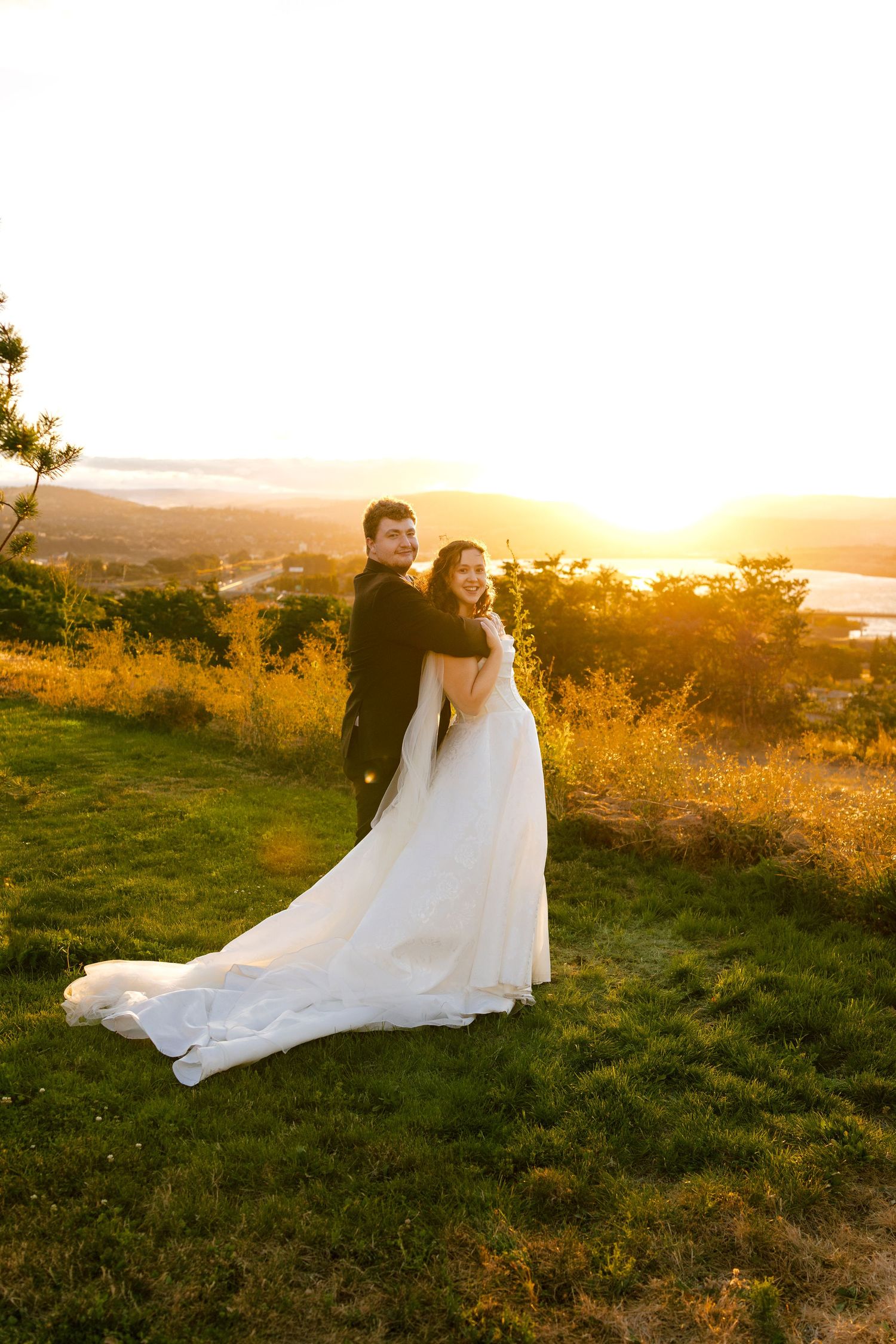 A couple in wedding attire embraces during golden hour sunset on a grassy hilltop overlooking a scenic landscape.