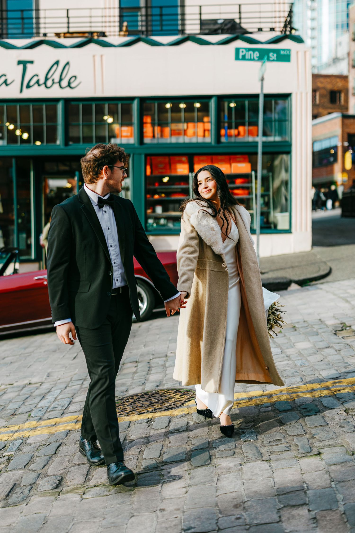Couple walking together on cobblestone street near urban storefronts.