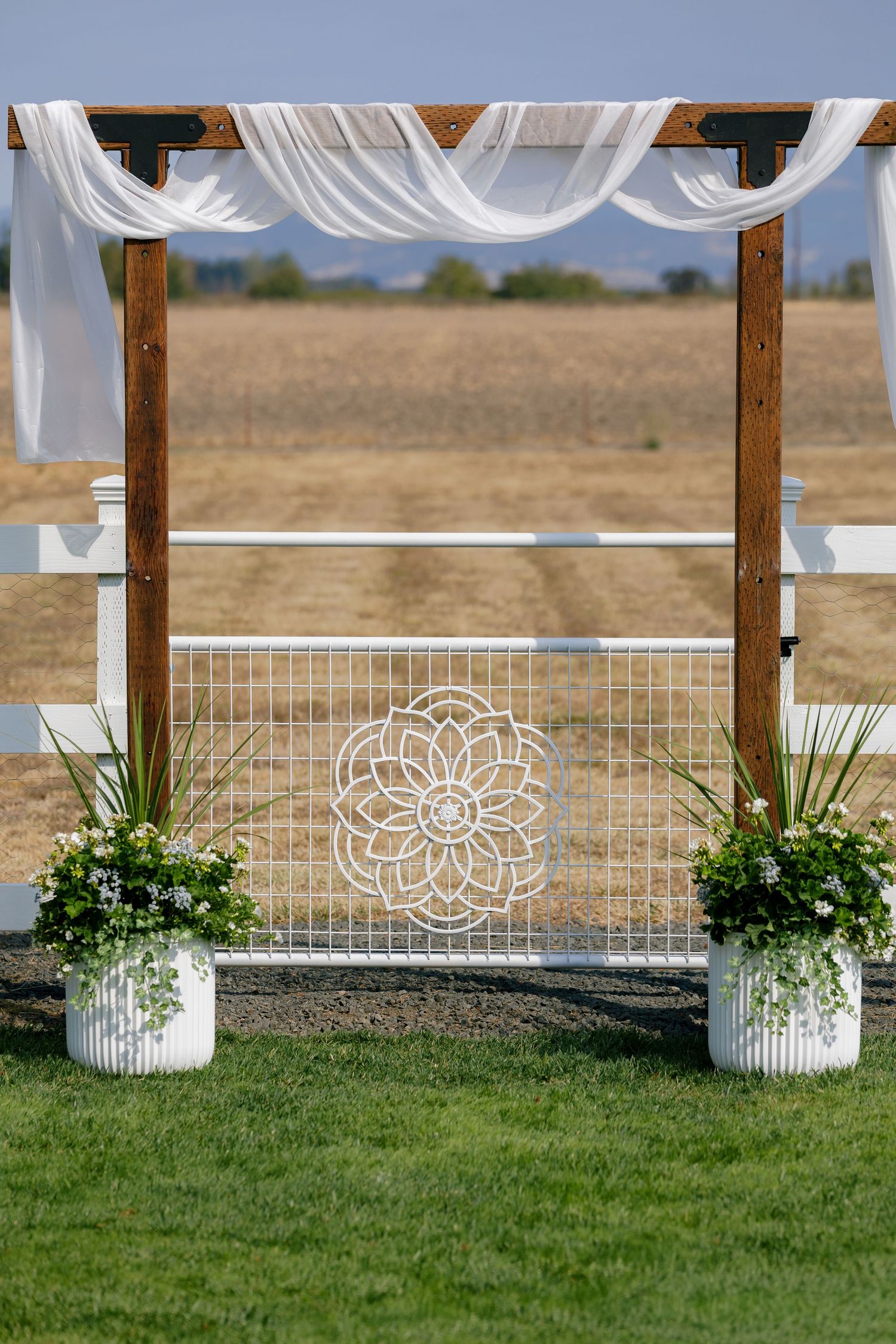 White wedding arch decorated with floral arrangements and fabric draping stands against a rural landscape.