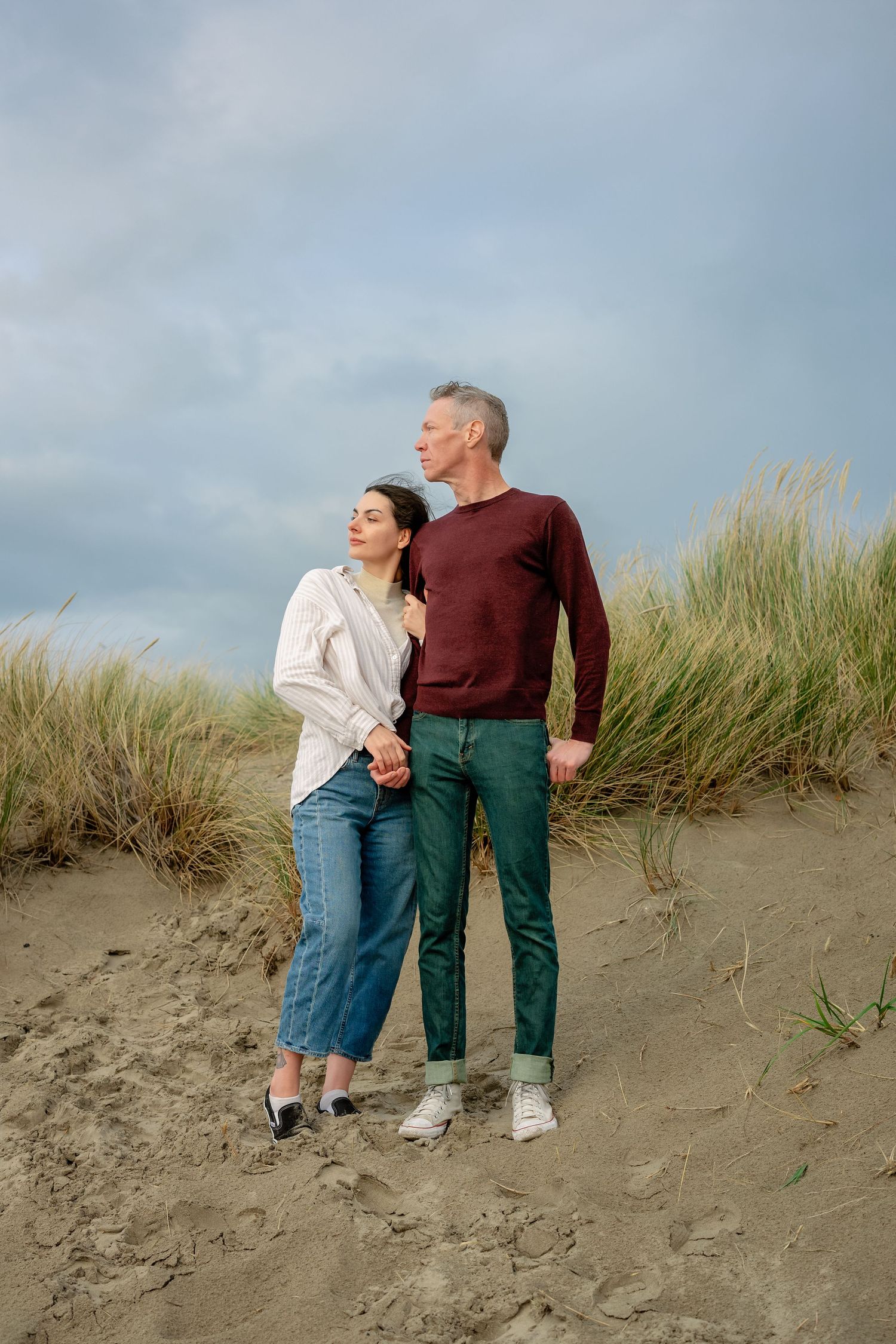 Two people in casual attire stand together on a sandy beach dune with tall grass swaying in the background.