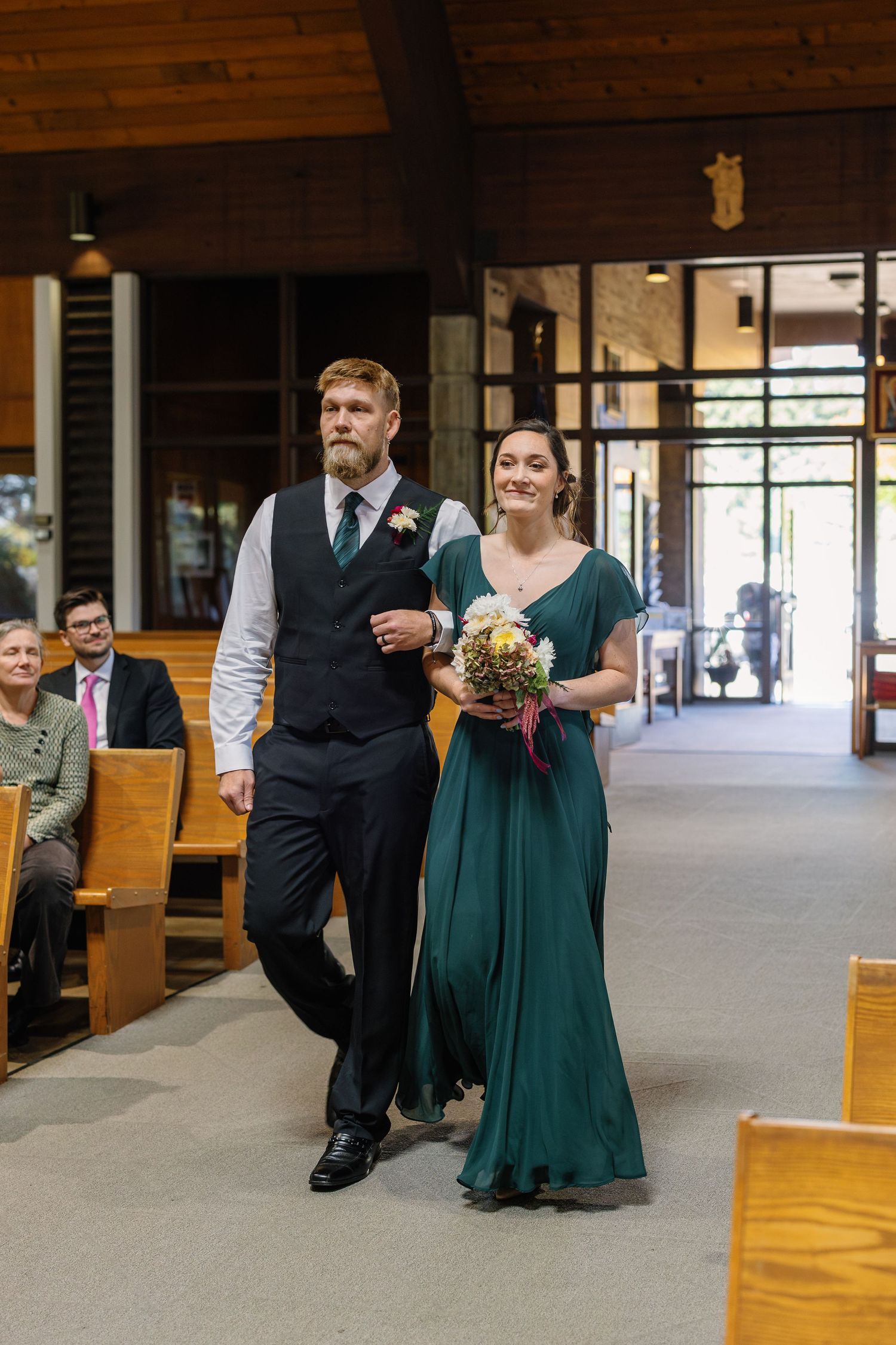 Wedding party members in emerald green dresses and dark suits walk down an aisle in a church.